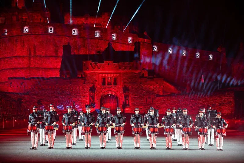 A marching band with drummers standing in formation on a stage, with a historic red stone fortress illuminated at night in the background, and dramatic lighting overhead.