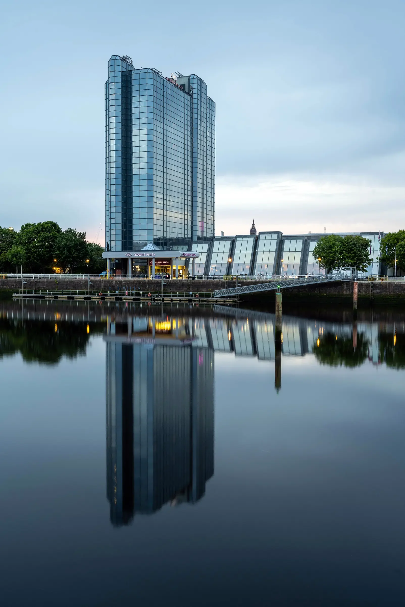 A tall, modern glass building by the water with its reflection visible in the calm river, overcast sky, and some greenery around.