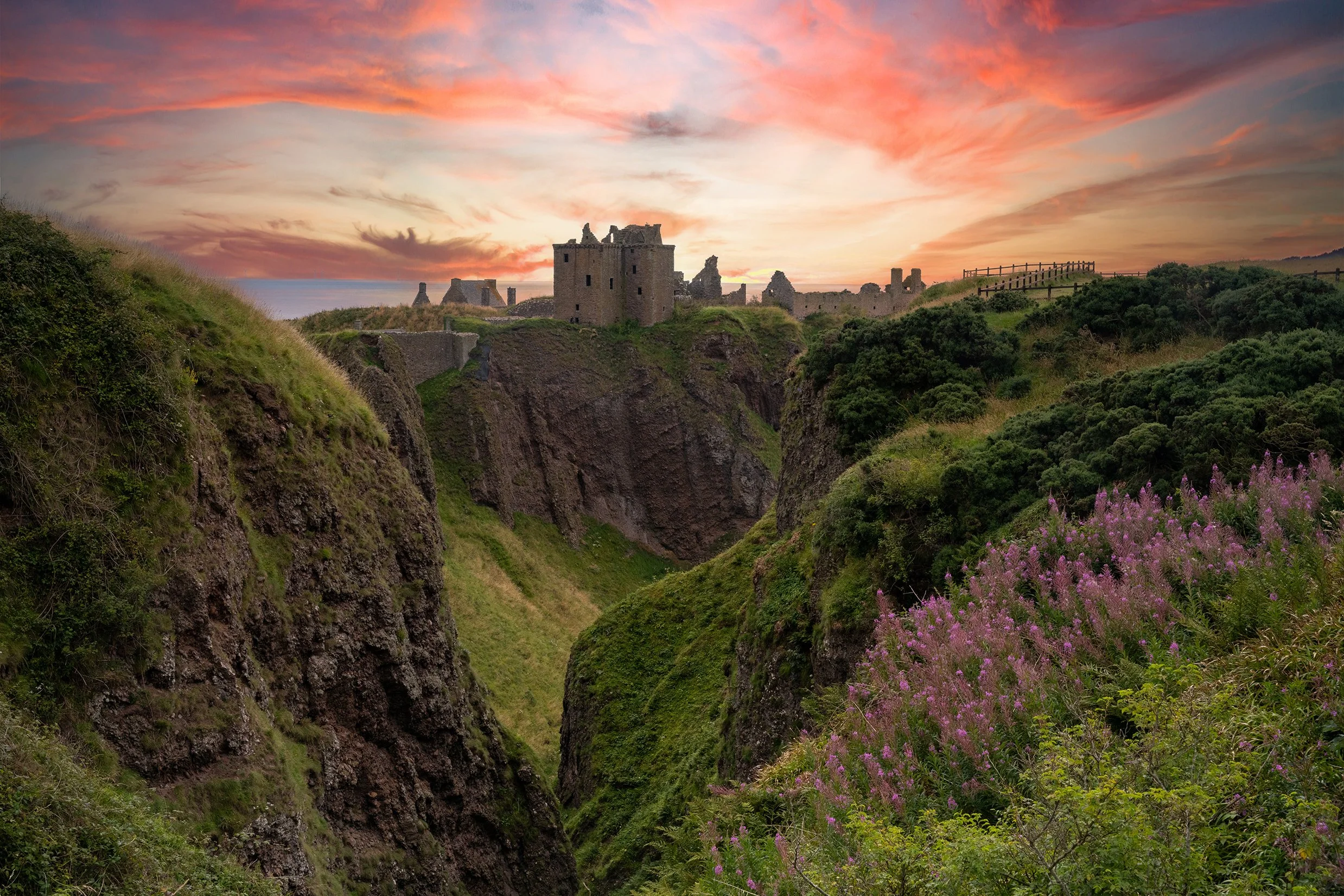 A scenic view of an ancient castle on a cliff during sunset, with pink and orange sky, lush greenery, purple wildflowers, and a wooden fence in the background.