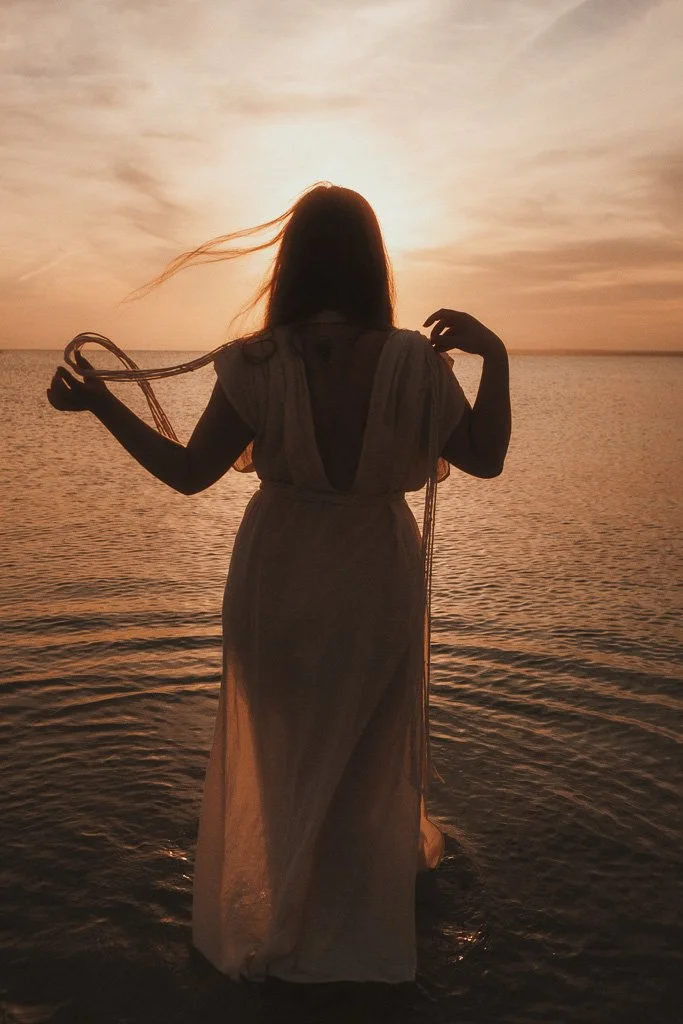 A woman standing in shallow water at sunset, wearing a long, flowing dress, with her hair blowing in the breeze.