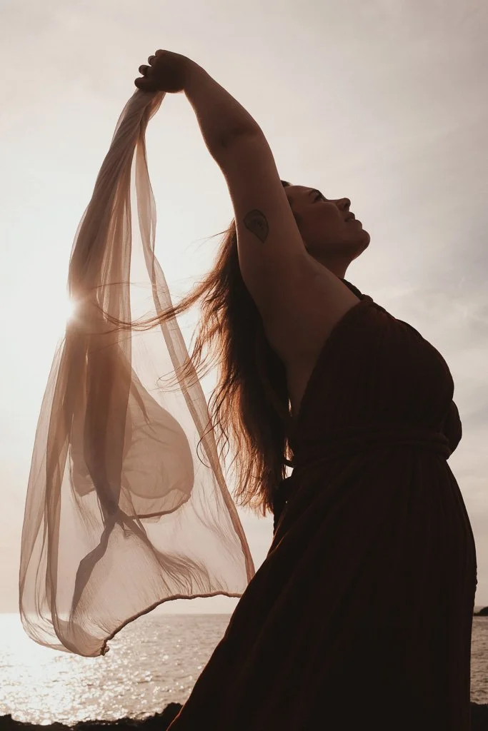 Frau beim Sonnenuntergang am Strand, hält einen transparenten Schal in der Luft.