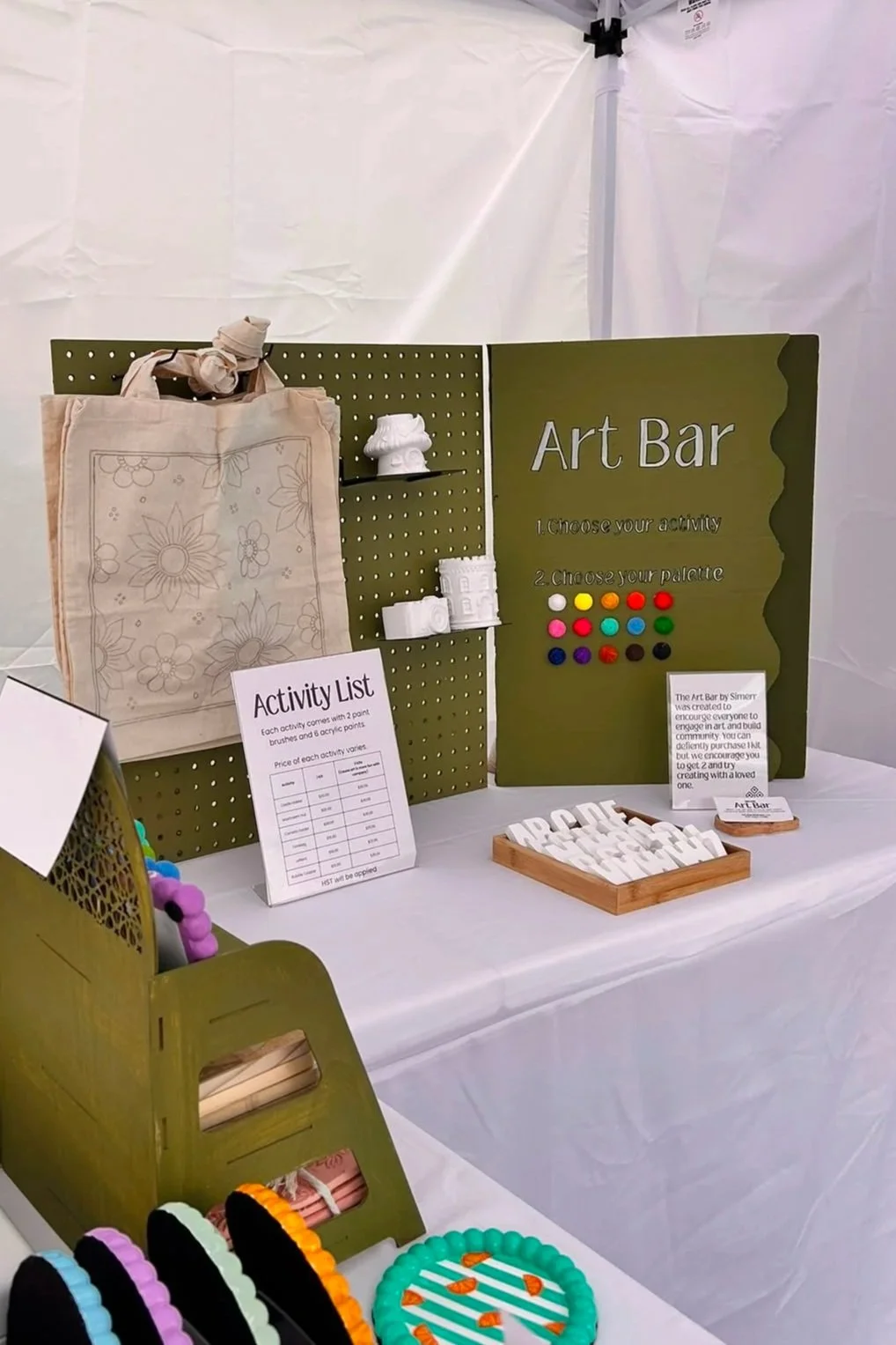 Sim.err mobile Art Bar setup at a market with tote bags, ceramic pieces, art activities and painting supplies.