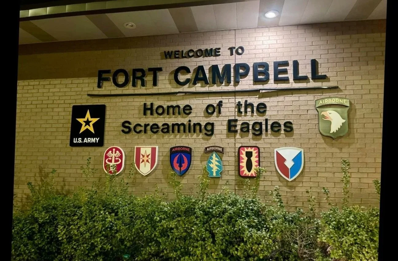 Wall with signs and emblems at Fort Campbell, including the U.S. Army star and aircraft badge, welcoming visitors with the text "Welcome to Fort Campbell, Home of the Screaming Eagles," and surrounded by bushes.