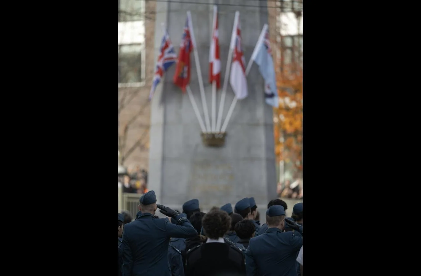 A group of uniformed individuals, likely military or veterans, saluting in front of a memorial with multiple flags, including the Union Jack and other British flags.