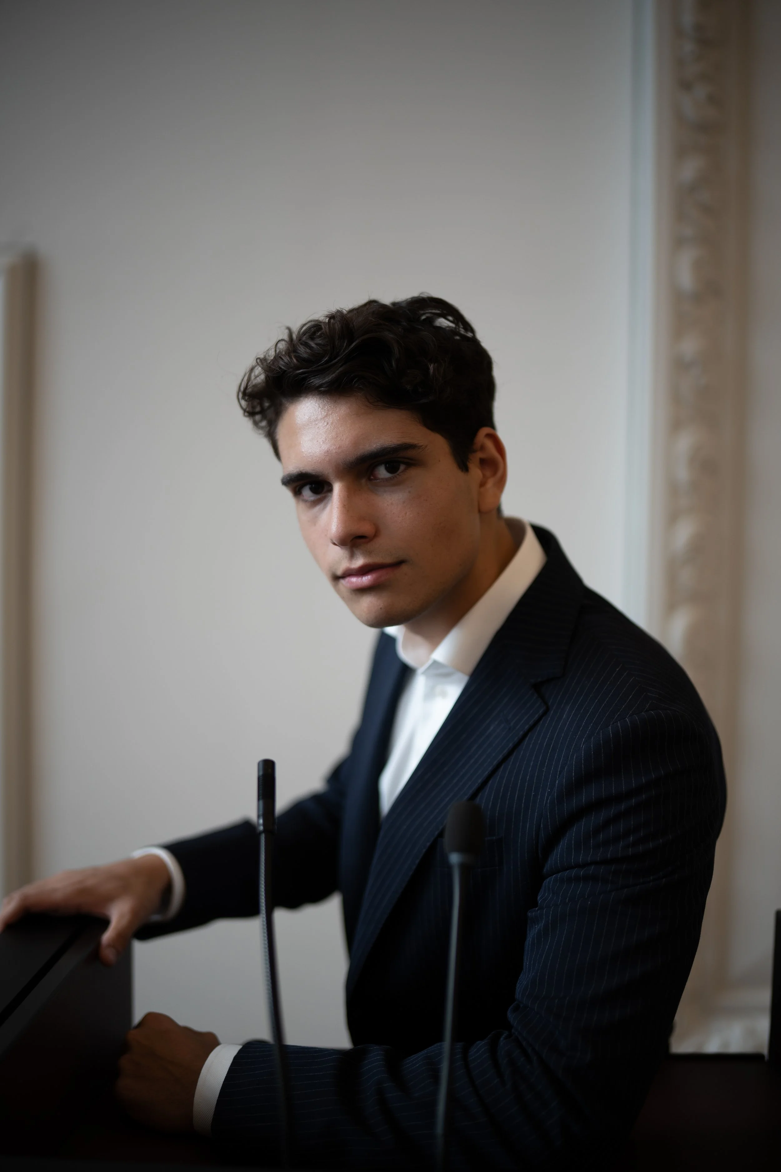 Young man in a dark pinstripe suit with white shirt, seated at a desk or table with microphones, looking into the camera.