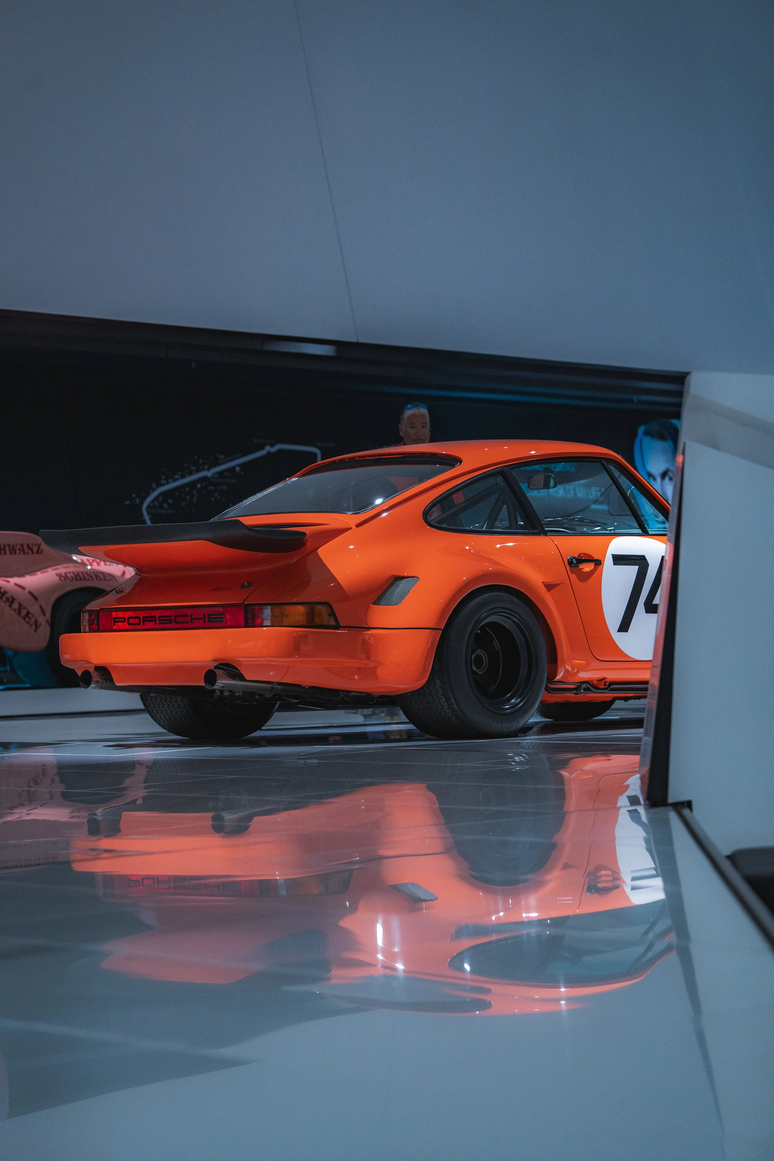 Orange Porsche race car displayed in a museum, with a reflection on the shiny floor and other cars partially visible nearby.