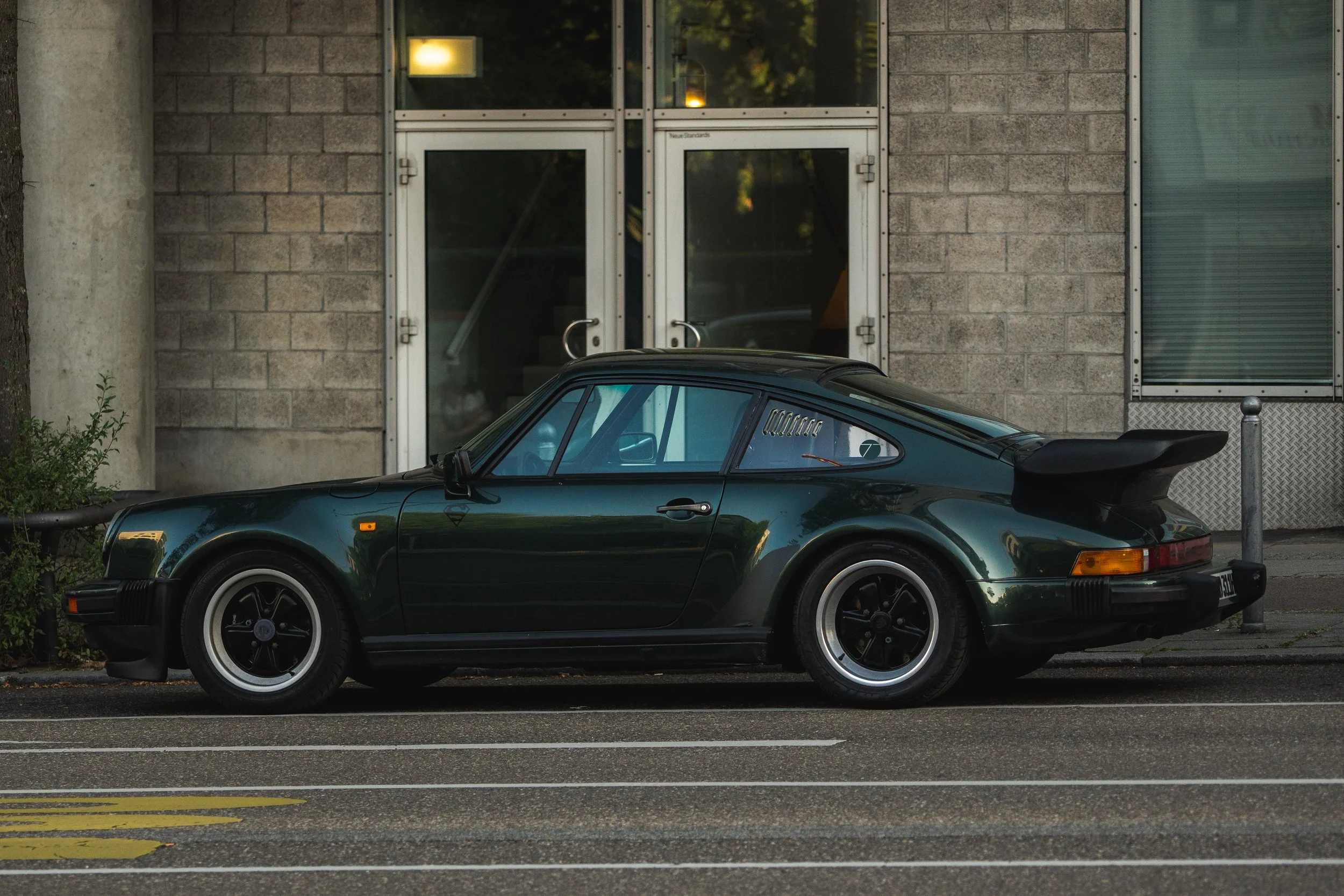 A dark green vintage Porsche 911 parked on the side of a city street in front of a building with concrete and glass doors.