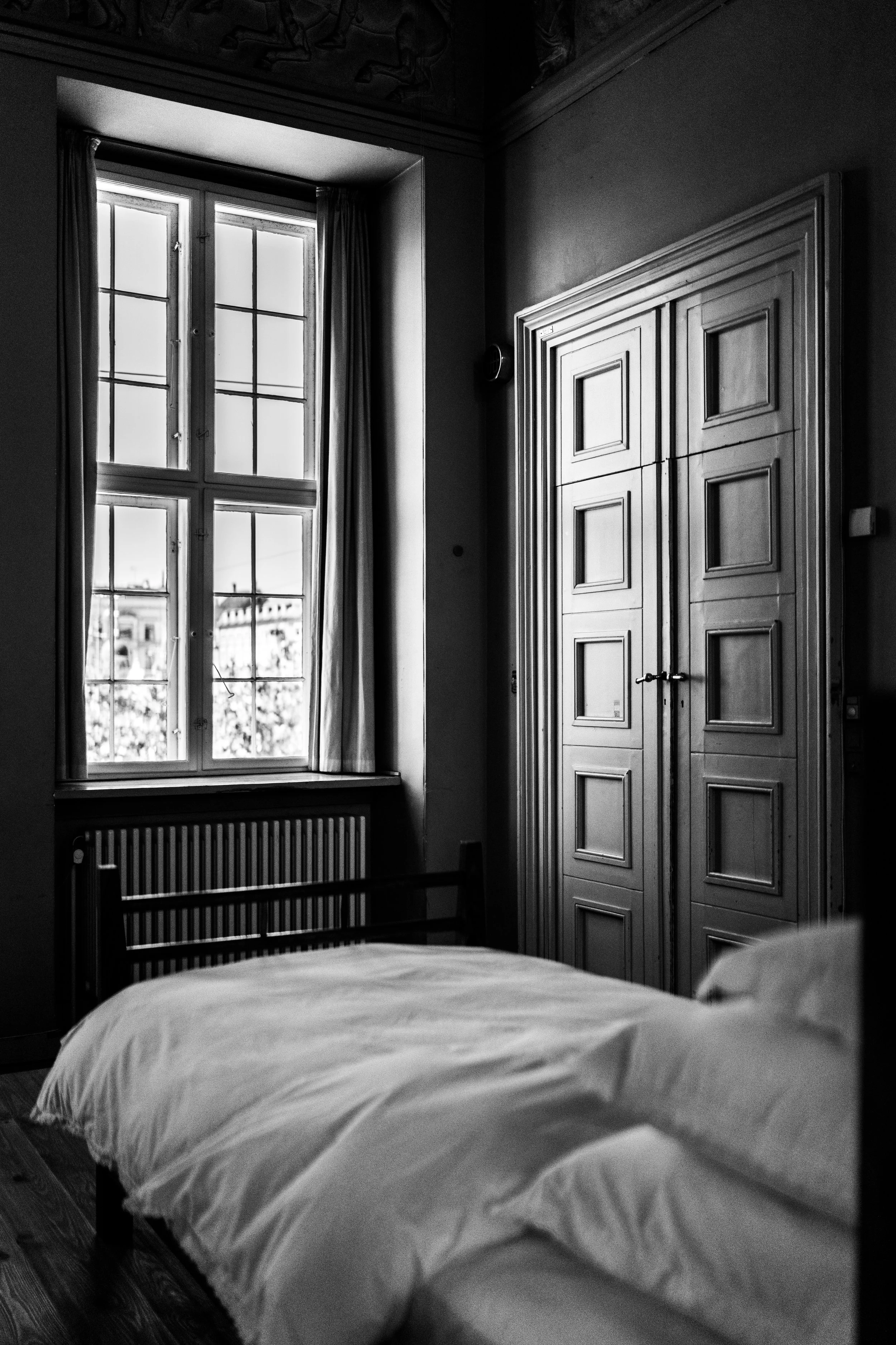 A black and white photo of a bedroom with a bed in the foreground, a window with curtains, and a large wooden wardrobe.