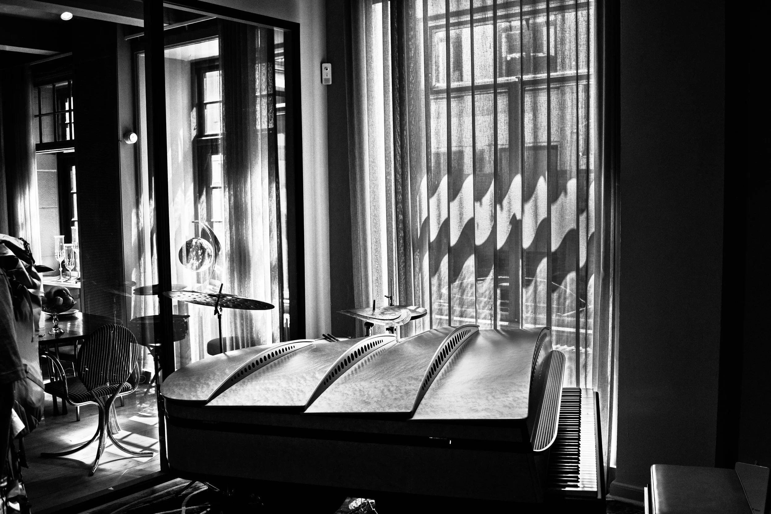 A black and white photo of a room with a modern piano on the right, sunlight coming through large windows with striped shadows, and a small drum set in the background.