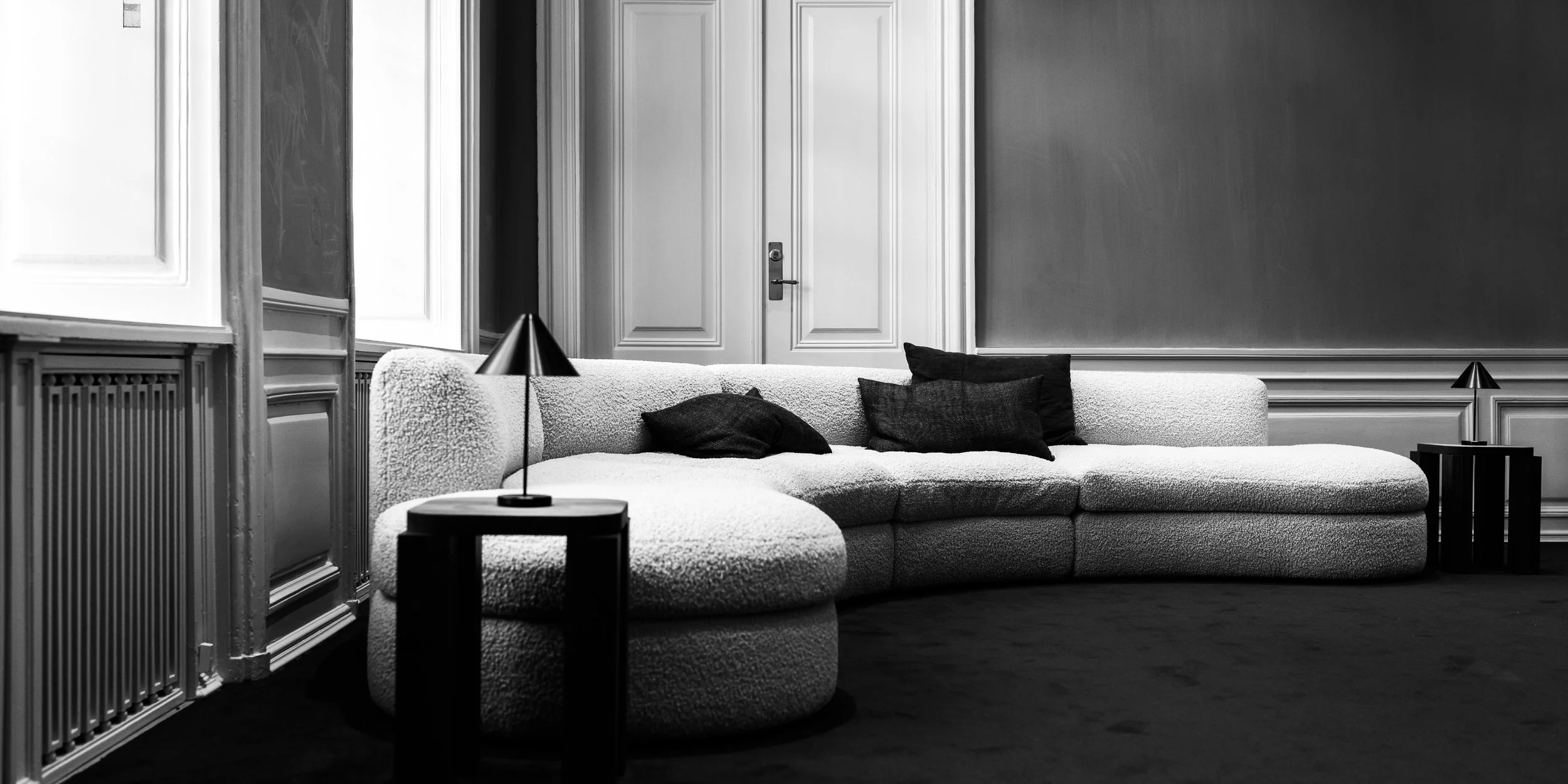A black and white photo of a modern living room with a textured sofa, darker pillows, and two side tables with small lamps. A door is visible in the background.