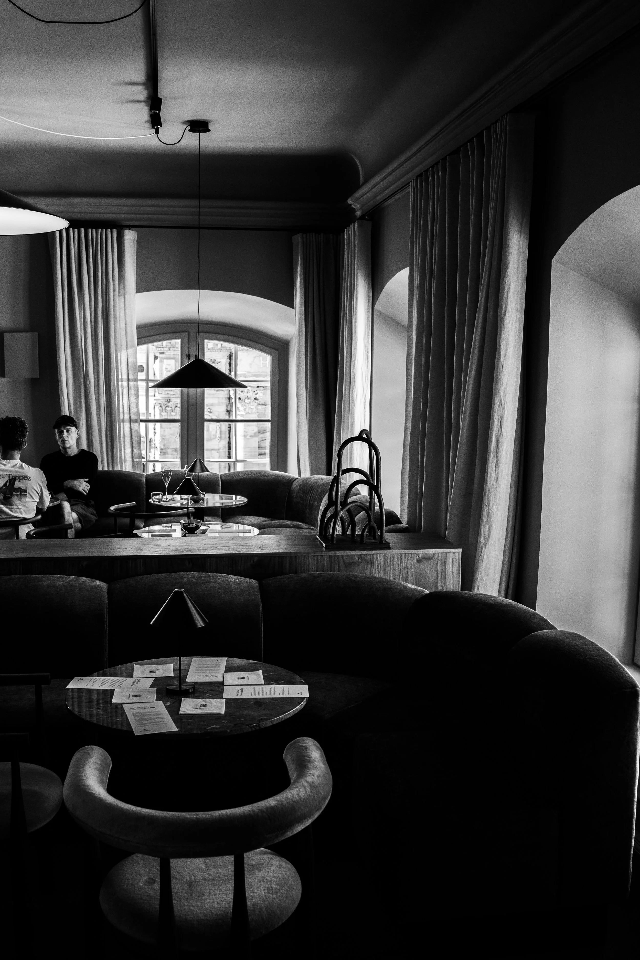 A black and white photo of a cozy restaurant interior with large windows, curtains, tables, and chairs, with two people seated in the background.