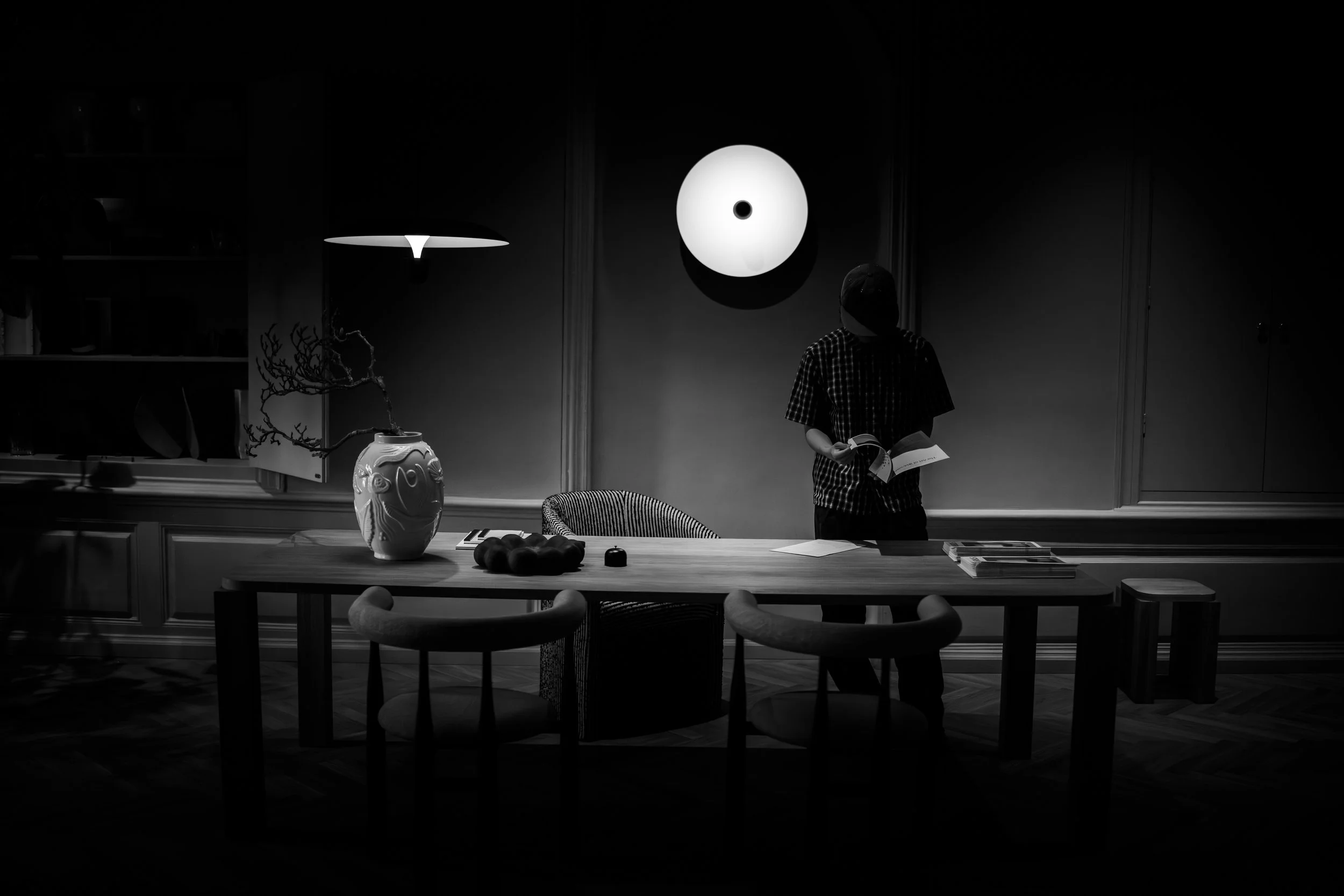 A person standing at a table reading a book in a dimly lit room with modern decor, a large vase, a chair, and a circular wall art piece illuminated overhead.