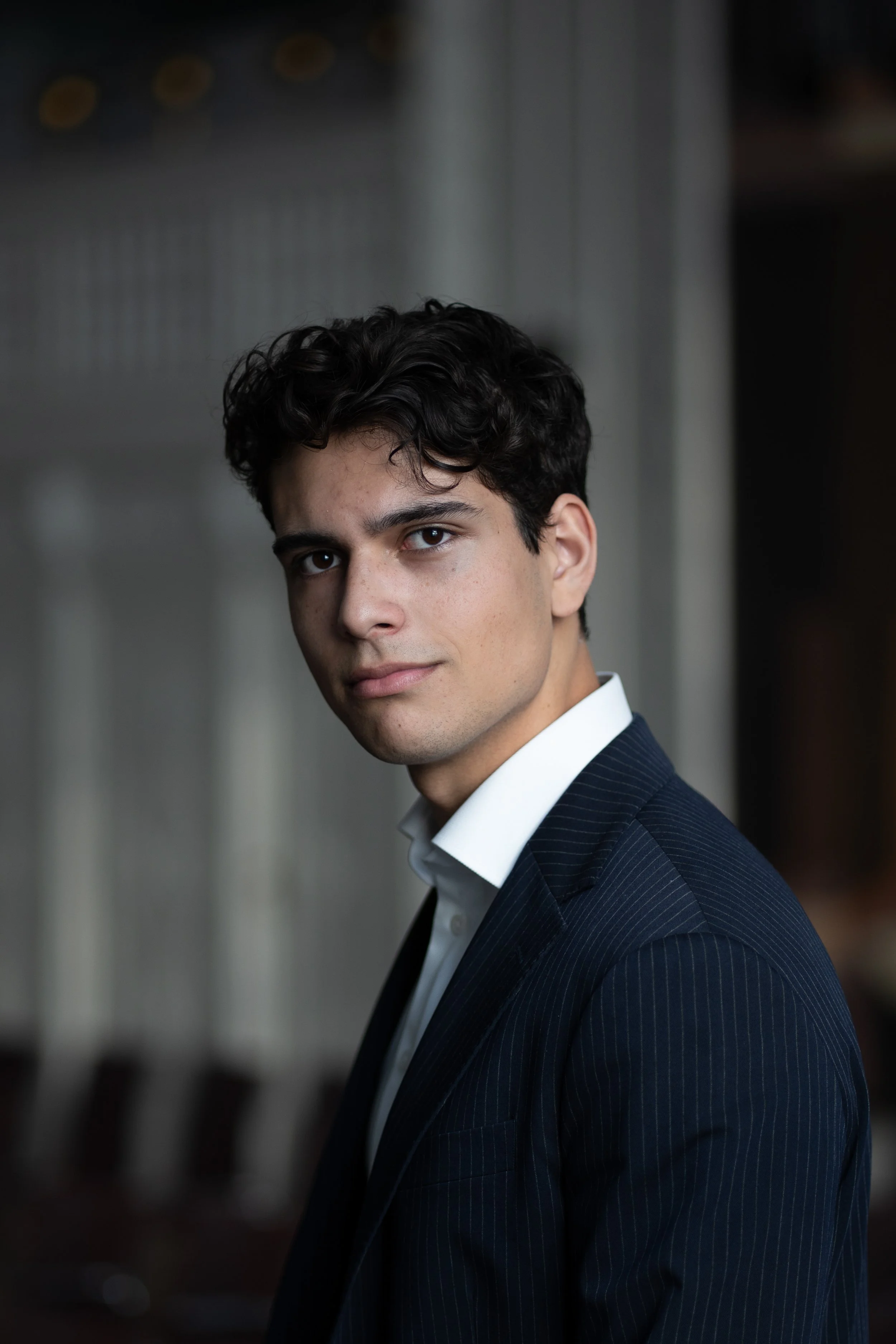 Portrait of a young man with dark curly hair wearing a dark pinstripe suit and white shirt, standing indoors with a blurred background.