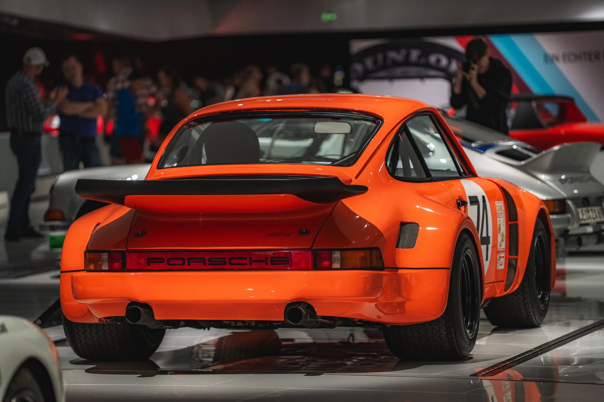 Rear view of an orange Porsche race car with a large black rear wing, parked indoors with other cars and people in the background.