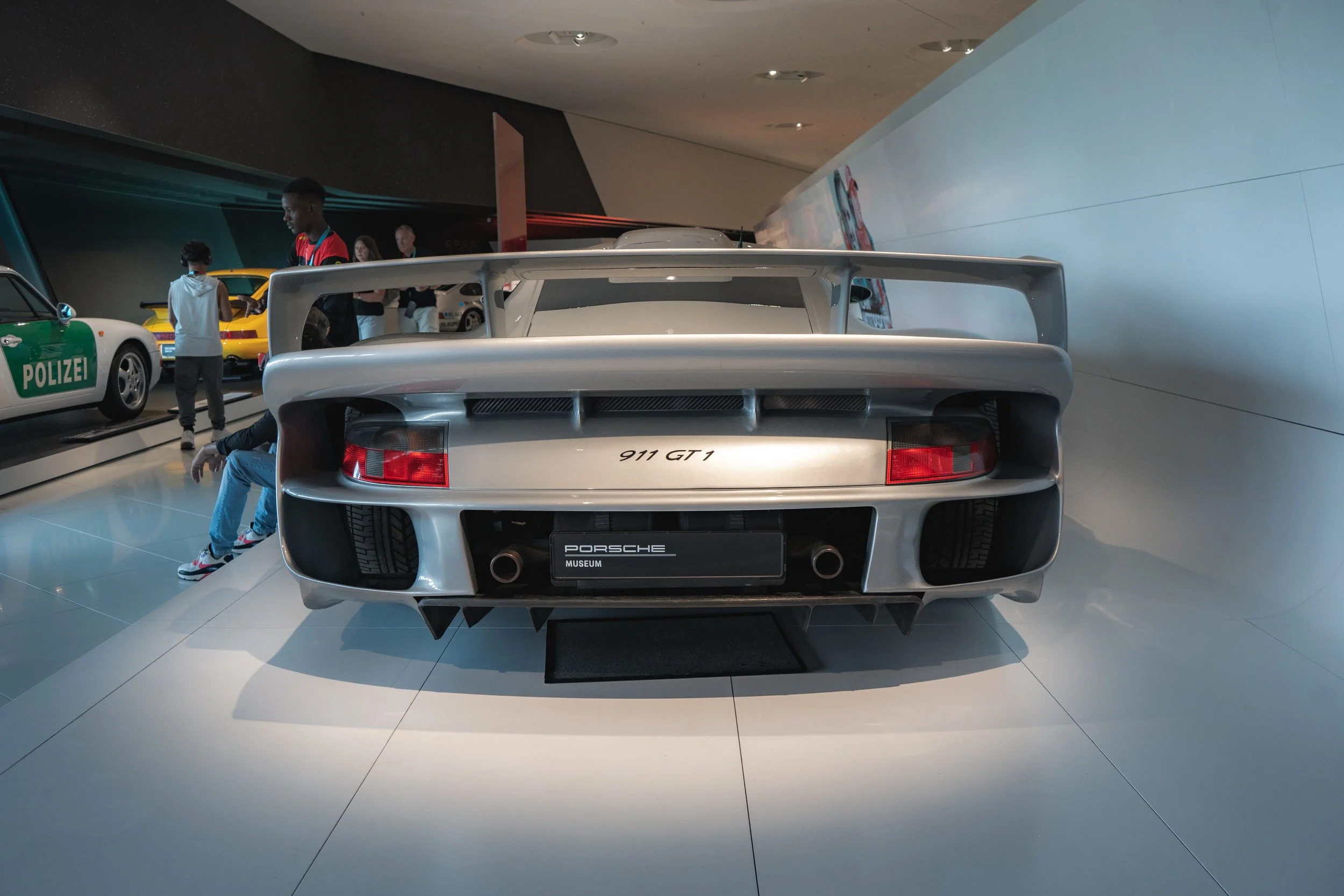 Rear view of a silver Porsche 911 GT1 displayed in a museum, with a black license plate and visible exhaust pipes, surrounded by other cars and visitors.