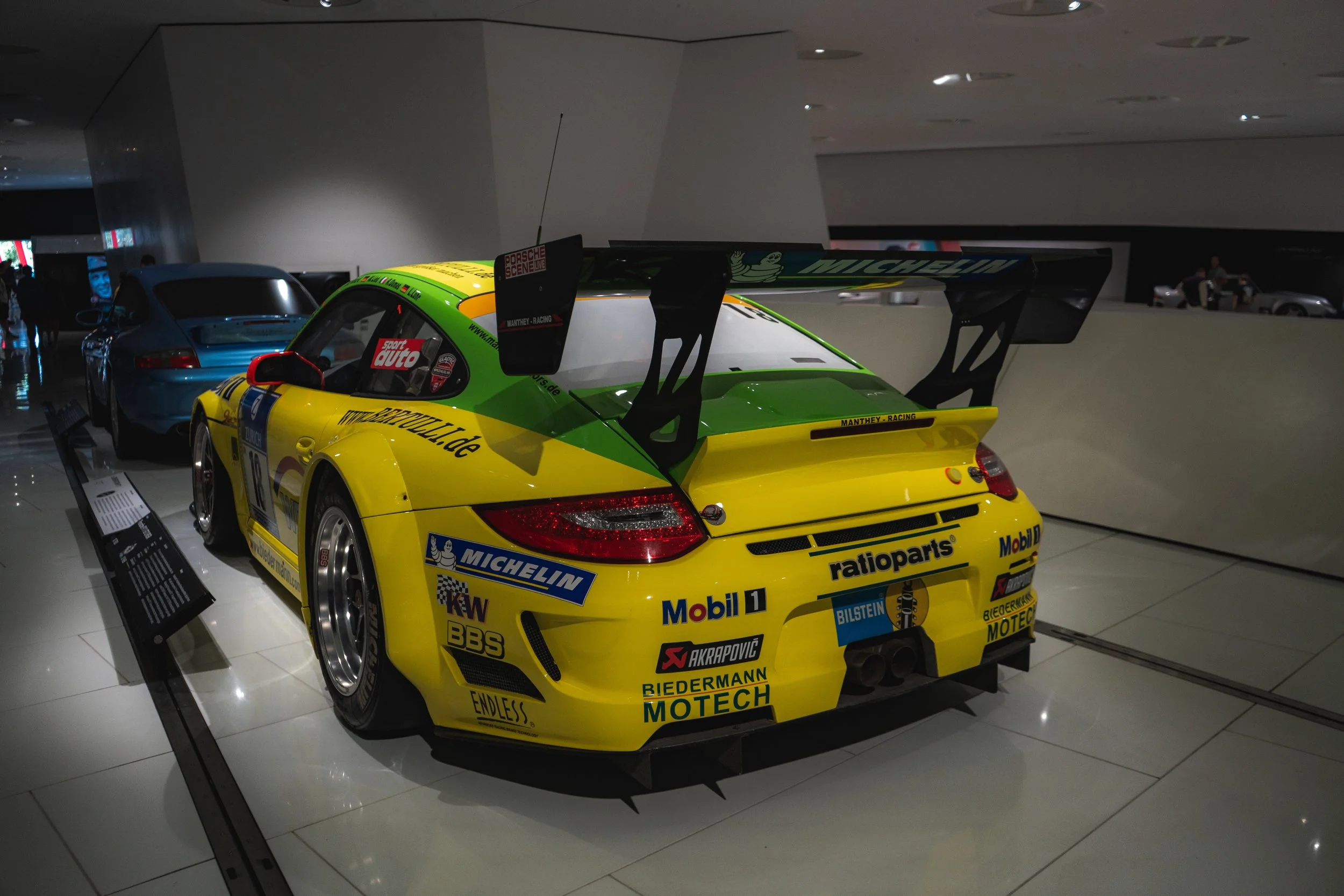Yellow and green racing car with sponsor decals on display inside a museum, with another car partially visible behind it.