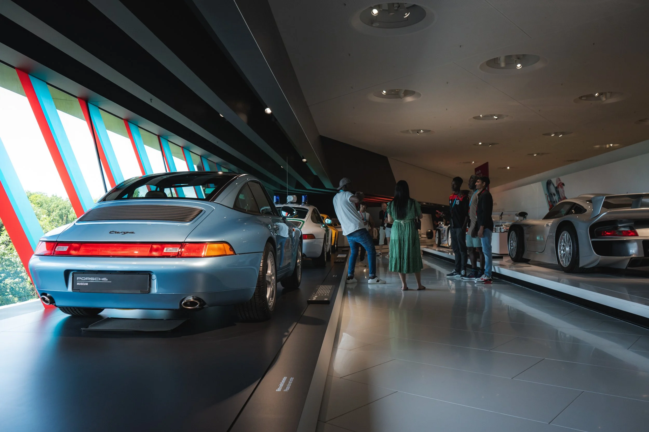 People viewing and discussing Porsche sports cars in an auto museum exhibit with large windows and colorful wall accents.