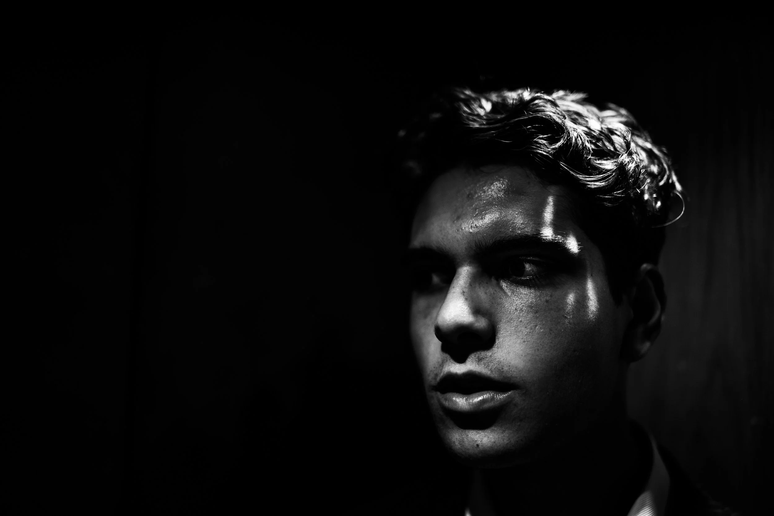 Black and white close-up portrait of a young man with curly hair, illuminated by a dramatic light source against a dark background.