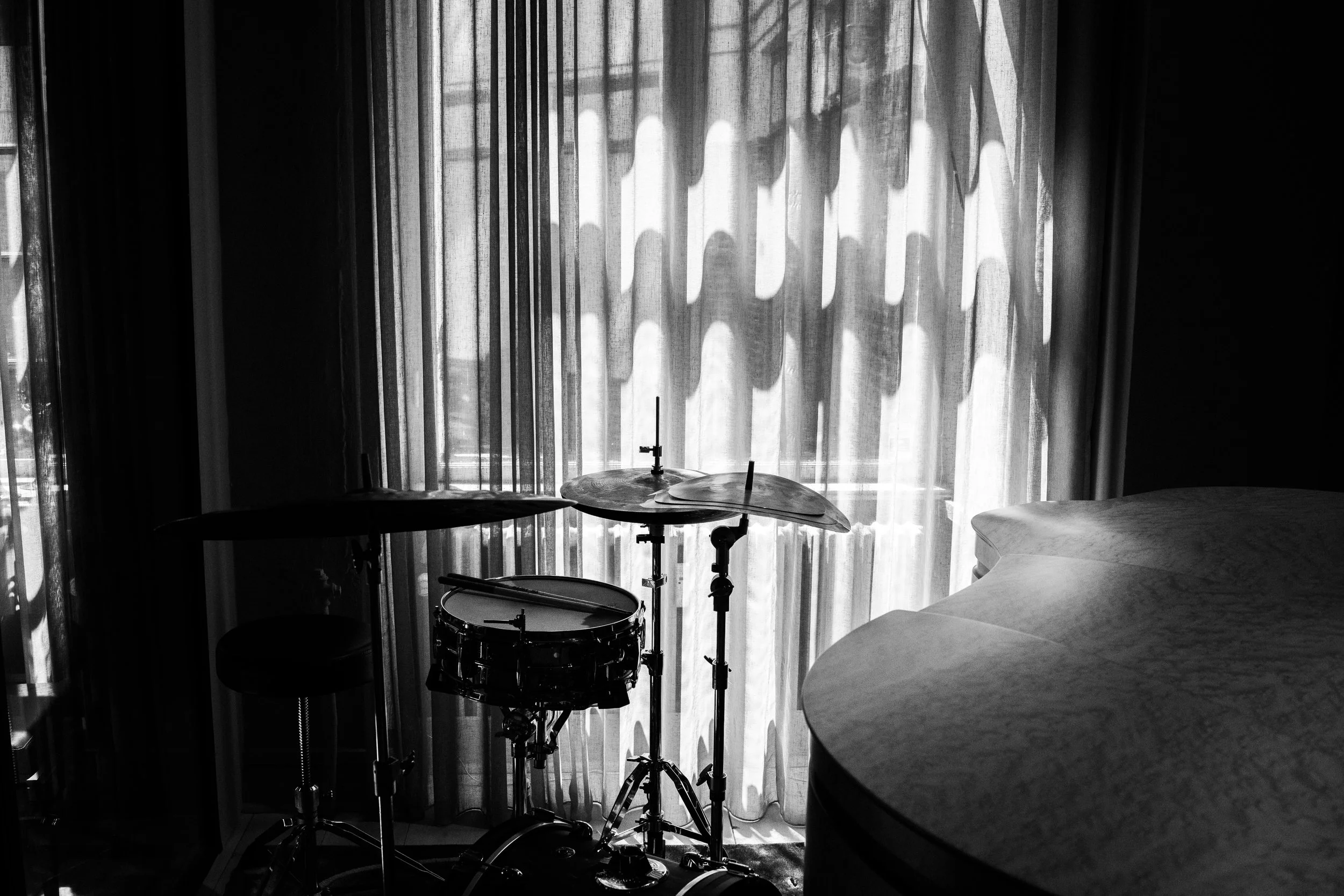 A drum set with a snare, cymbals, and a stool in front of a window with patterned curtains, in black and white.