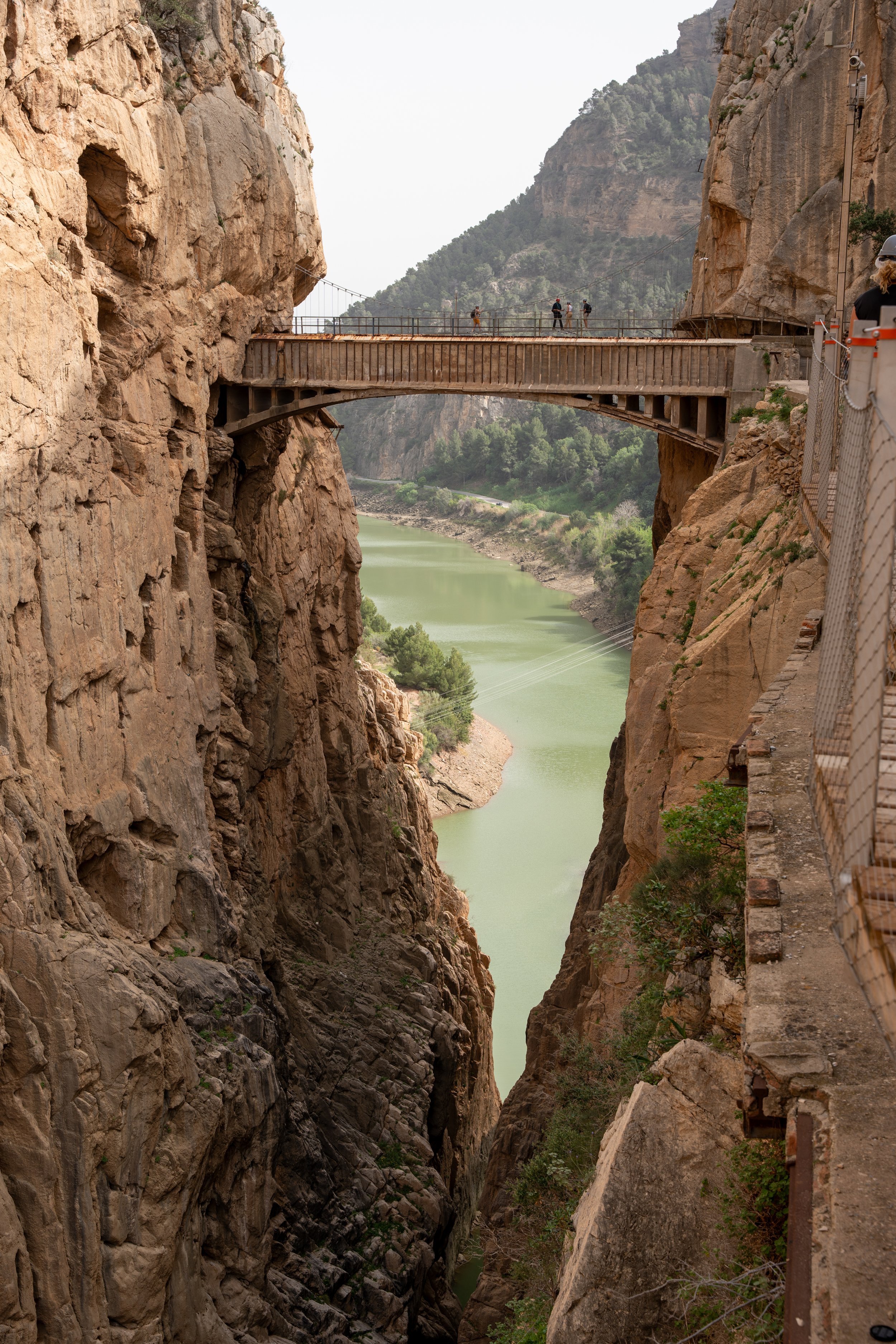 Walking trail along a narrow, rocky canyon with a river at the bottom, featuring a suspension bridge with hikers crossing.