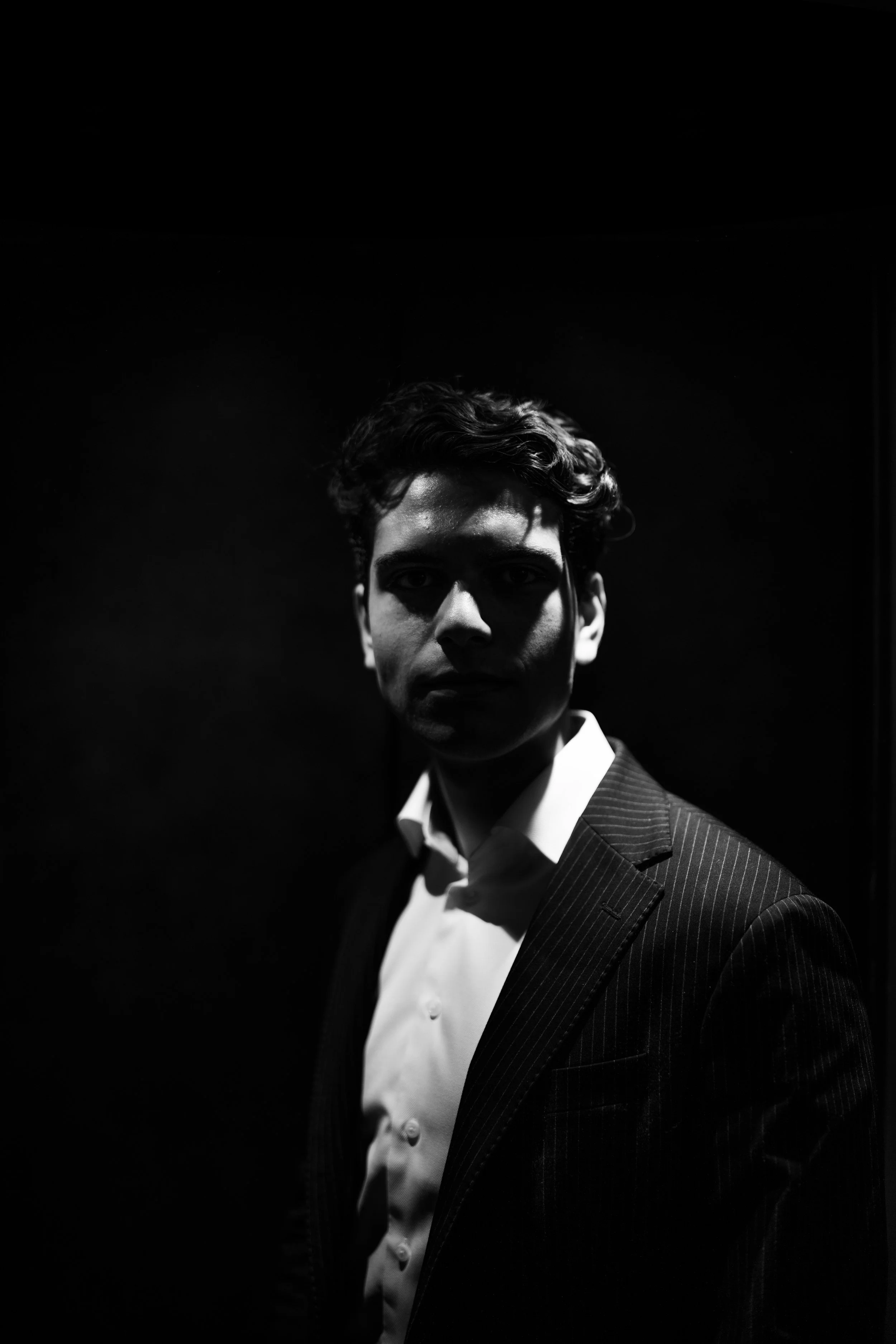 Black and white portrait of a young man with curly hair, wearing a suit and white shirt, standing in a dark setting with lighting emphasizing his face and upper body.