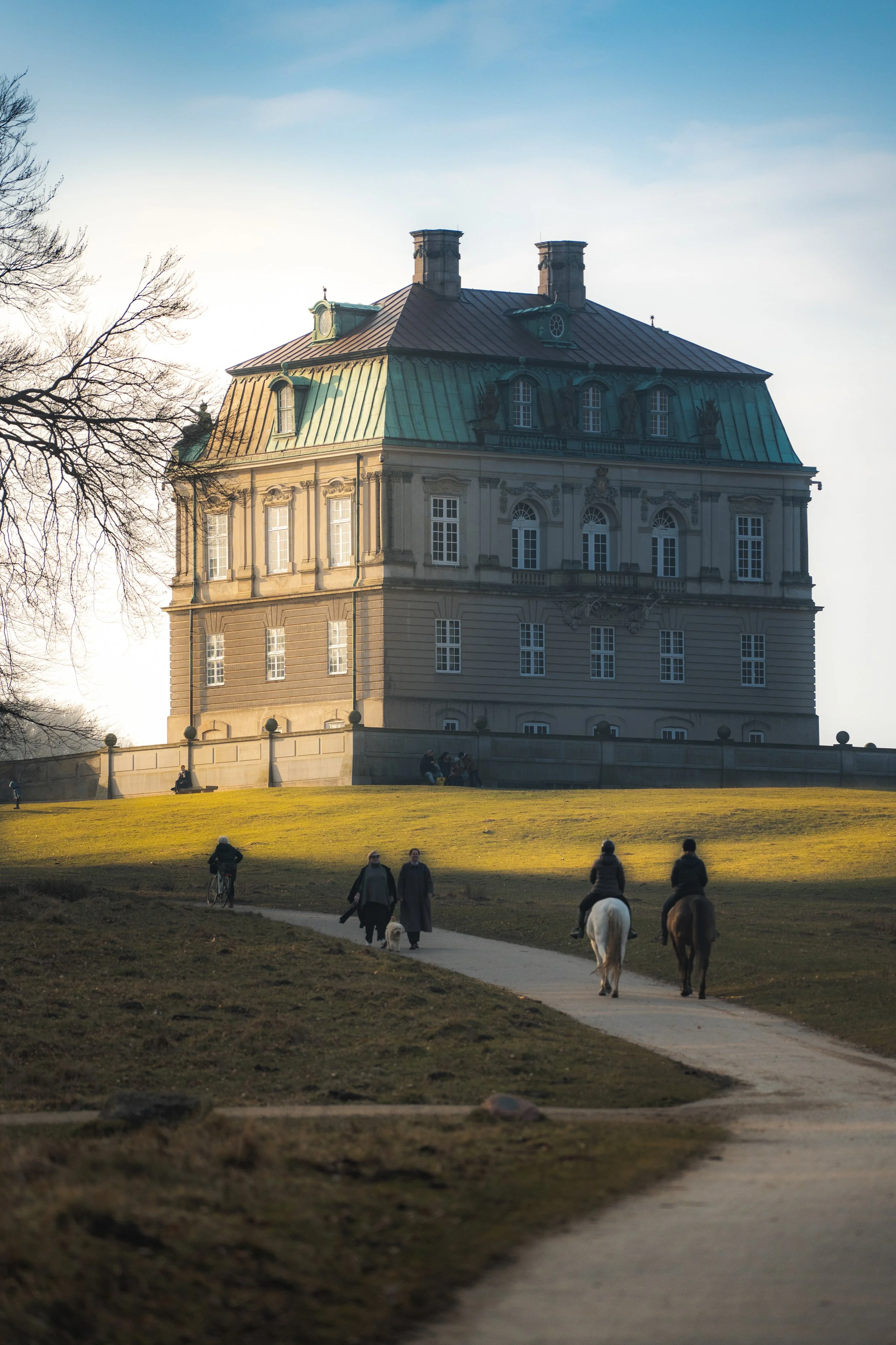Historical building with a green roof, situated on a grassy hill with people walking and riding horses in front of it.