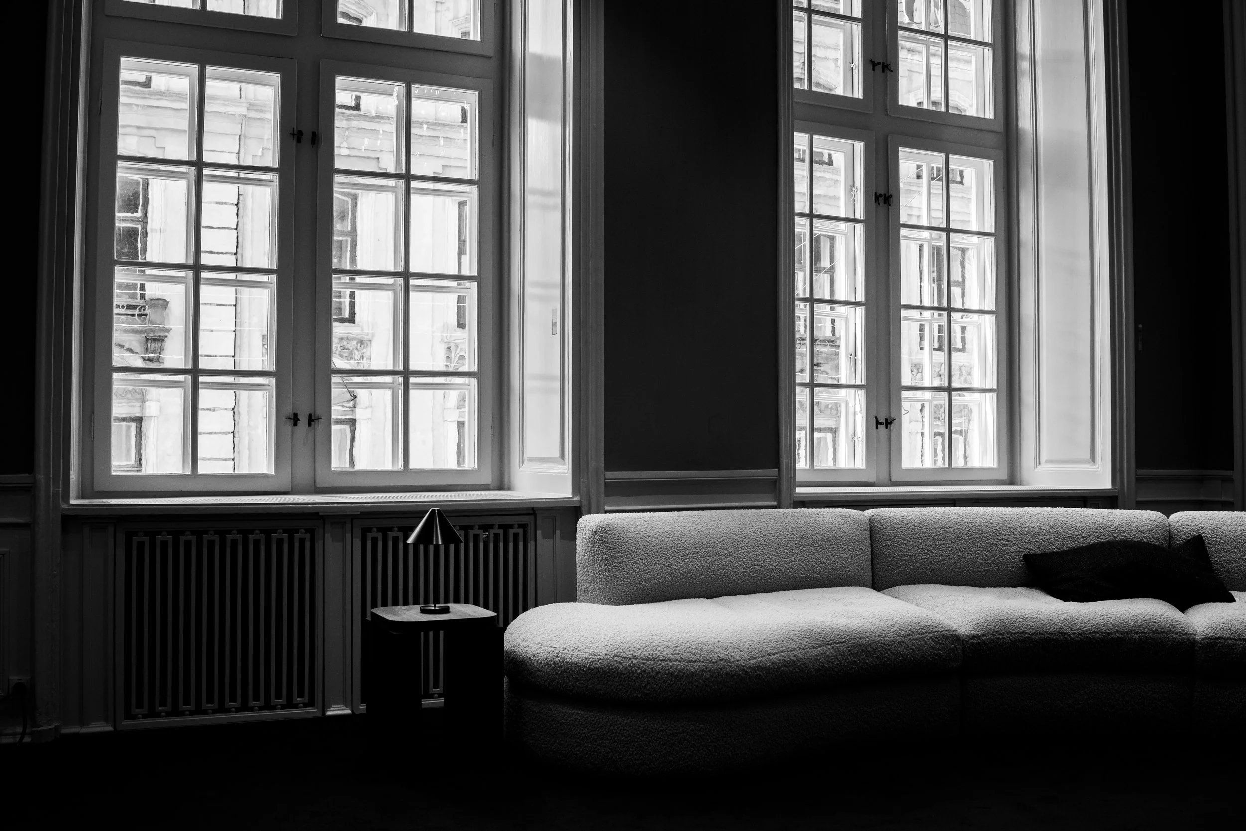 A black and white photo of a living room with three large windows. There is a small side table with a lamp, and a textured sofa with a cushion near the windows.