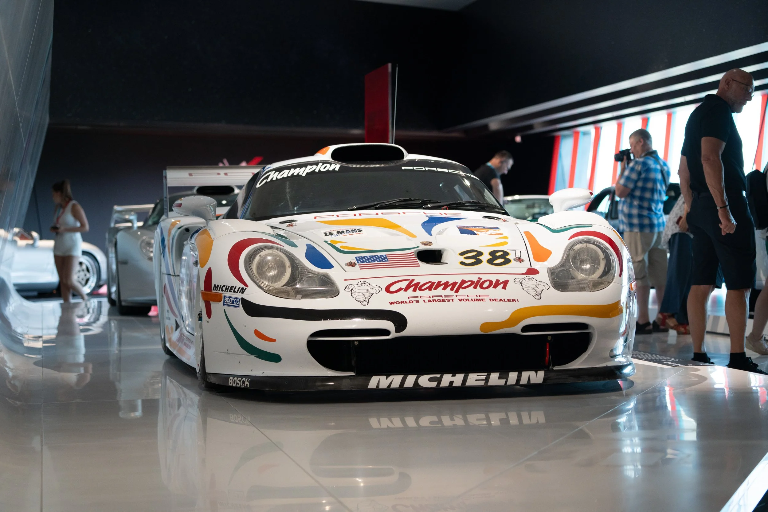 A white Porsche race car with colorful sponsor decals and the number 38 displayed, on display at an auto exhibit, with people walking and viewing in the background.