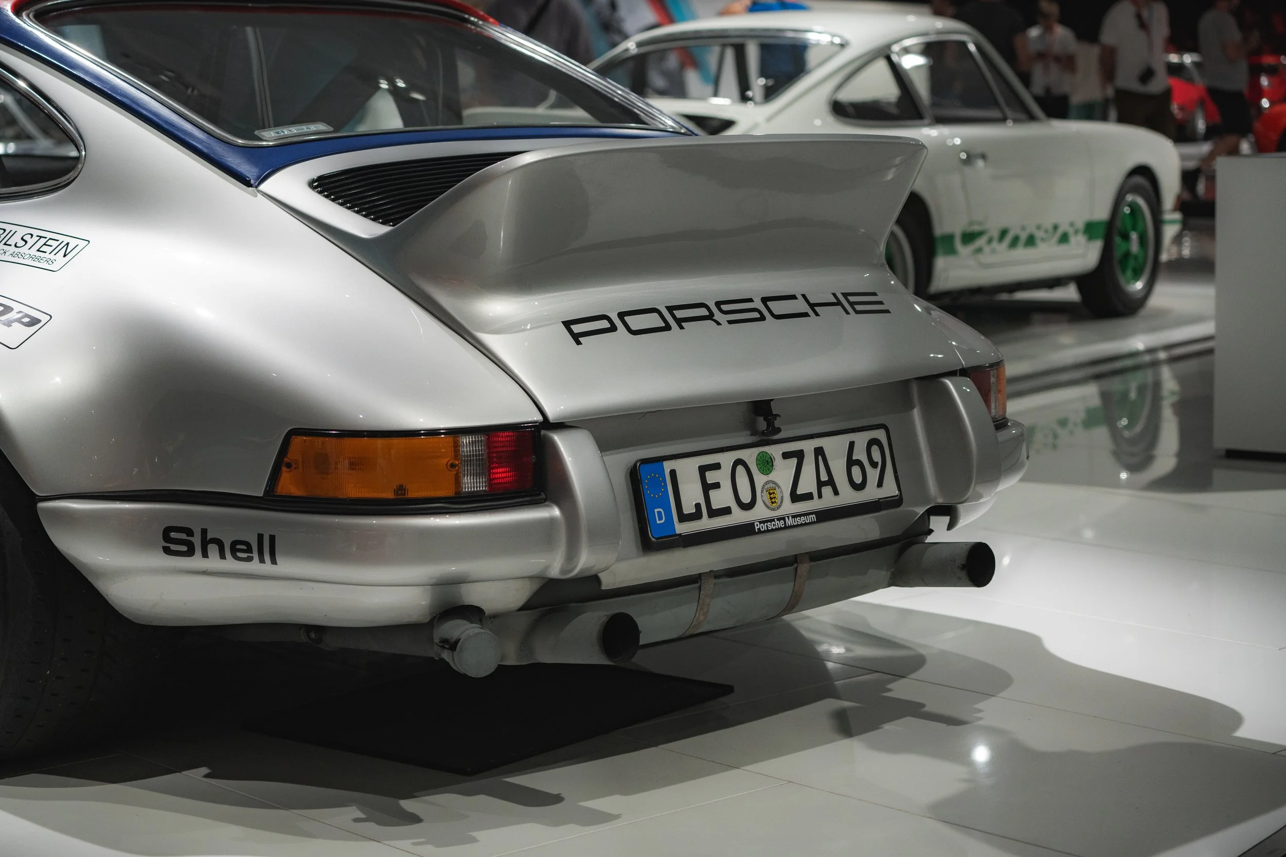 Rear view of a silver Porsche 911 racing car with a large rear wing, displaying Porsche logos and a German license plate marked 'LEO ZA 69', inside a car museum with another classic race car in the background.