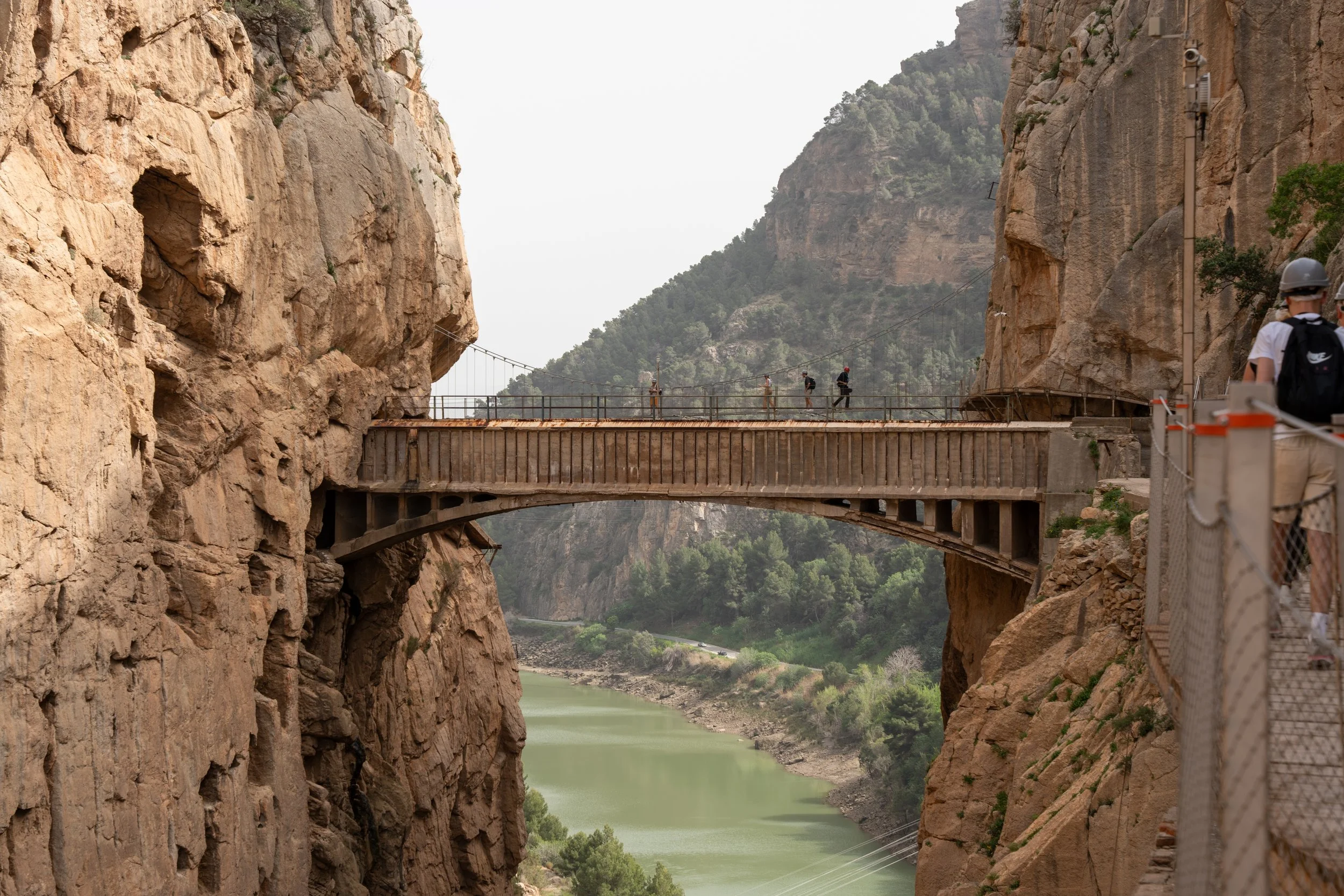 A walkway built across a deep canyon with rocky cliffs on both sides, a river flowing below, and green trees in the background. Several people are walking along the walkway.