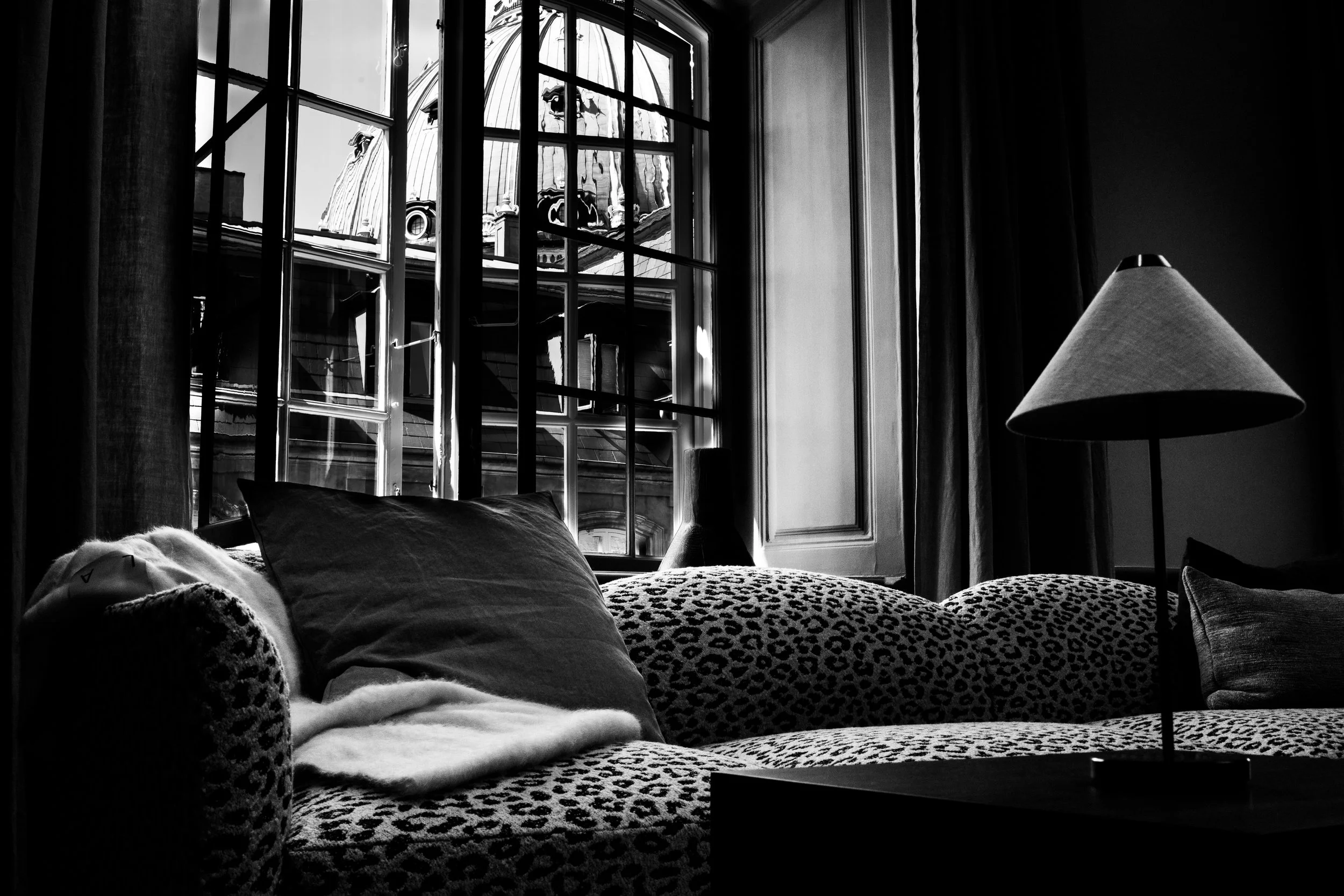 A black-and-white photo of a cozy living room with a patterned sofa, multiple pillows, a table lamp, and a large window showing a building with a dome on the roof outside.