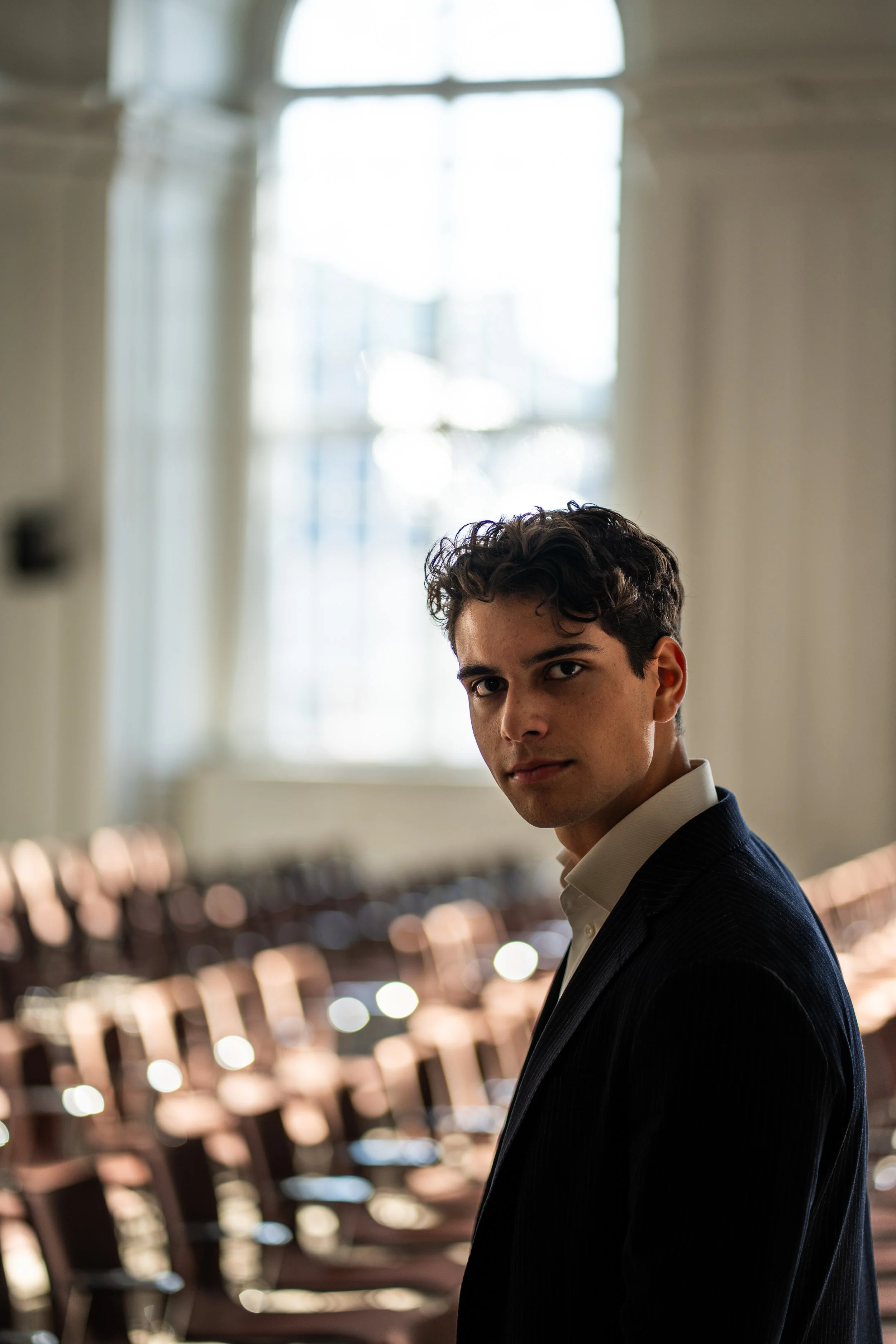 A young man in a dark suit and white shirt standing in a brightly lit room with large windows and rows of chairs.