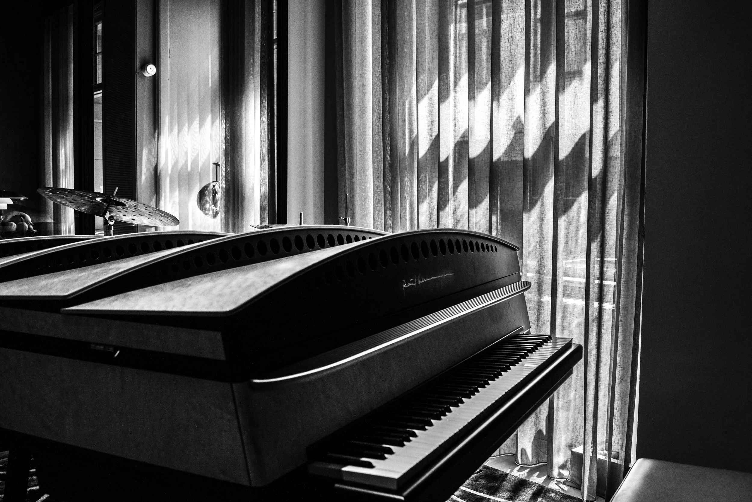 A black upright piano with a signature on the front, set next to a window with sheer curtains, sunlight casting patterns on the curtain, and a drum cymbal in the background.