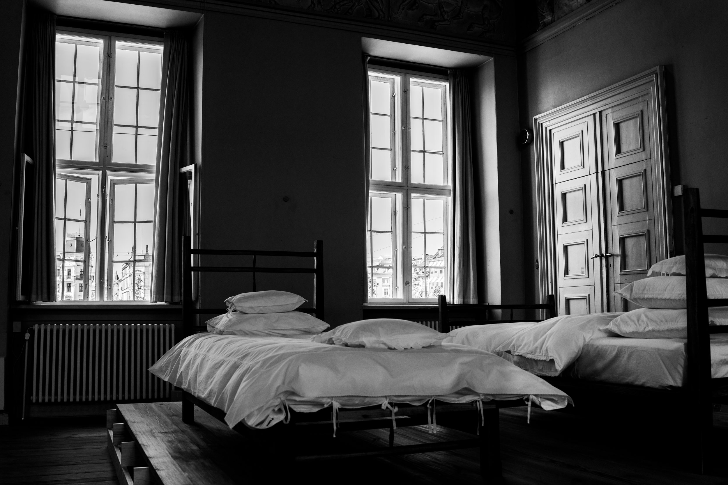 A black and white photo of a bedroom with two large windows, a bed with white bedding, and a tall wooden wardrobe.