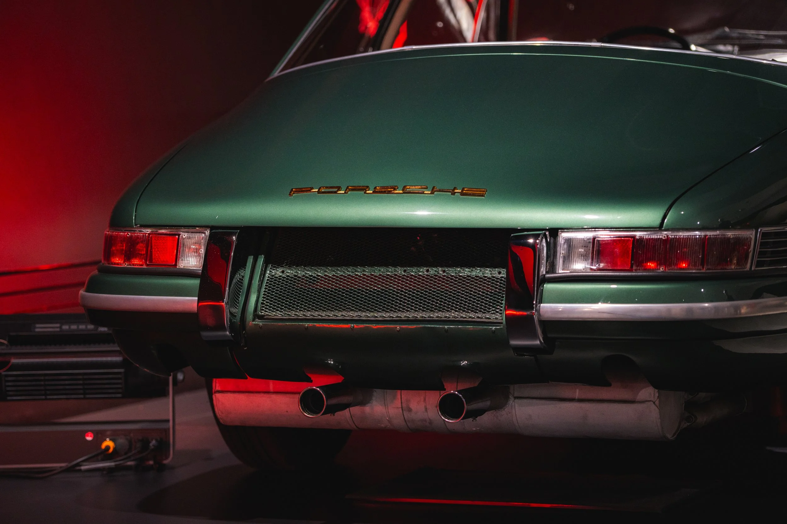 Close-up of a vintage green Porsche sports car rear, showing the Porsche emblem, taillights, dual exhaust pipes, and part of the rear bumper.