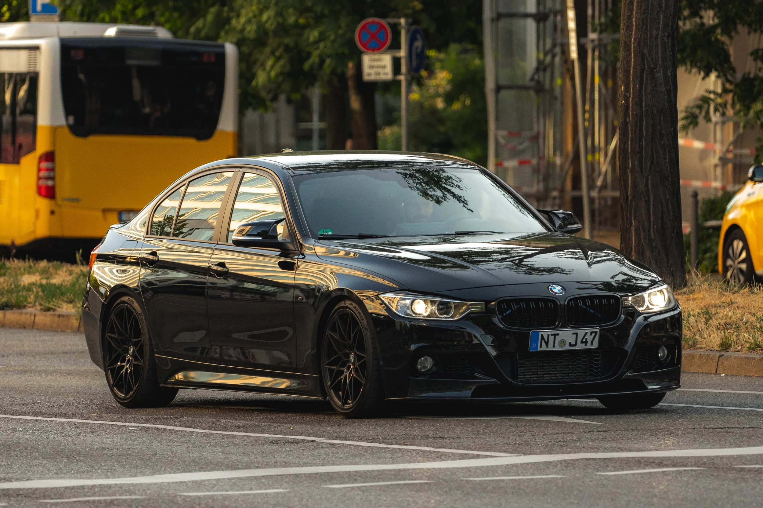 Black BMW sedan parked on city street at dusk with trees, a yellow bus, and signage in the background.