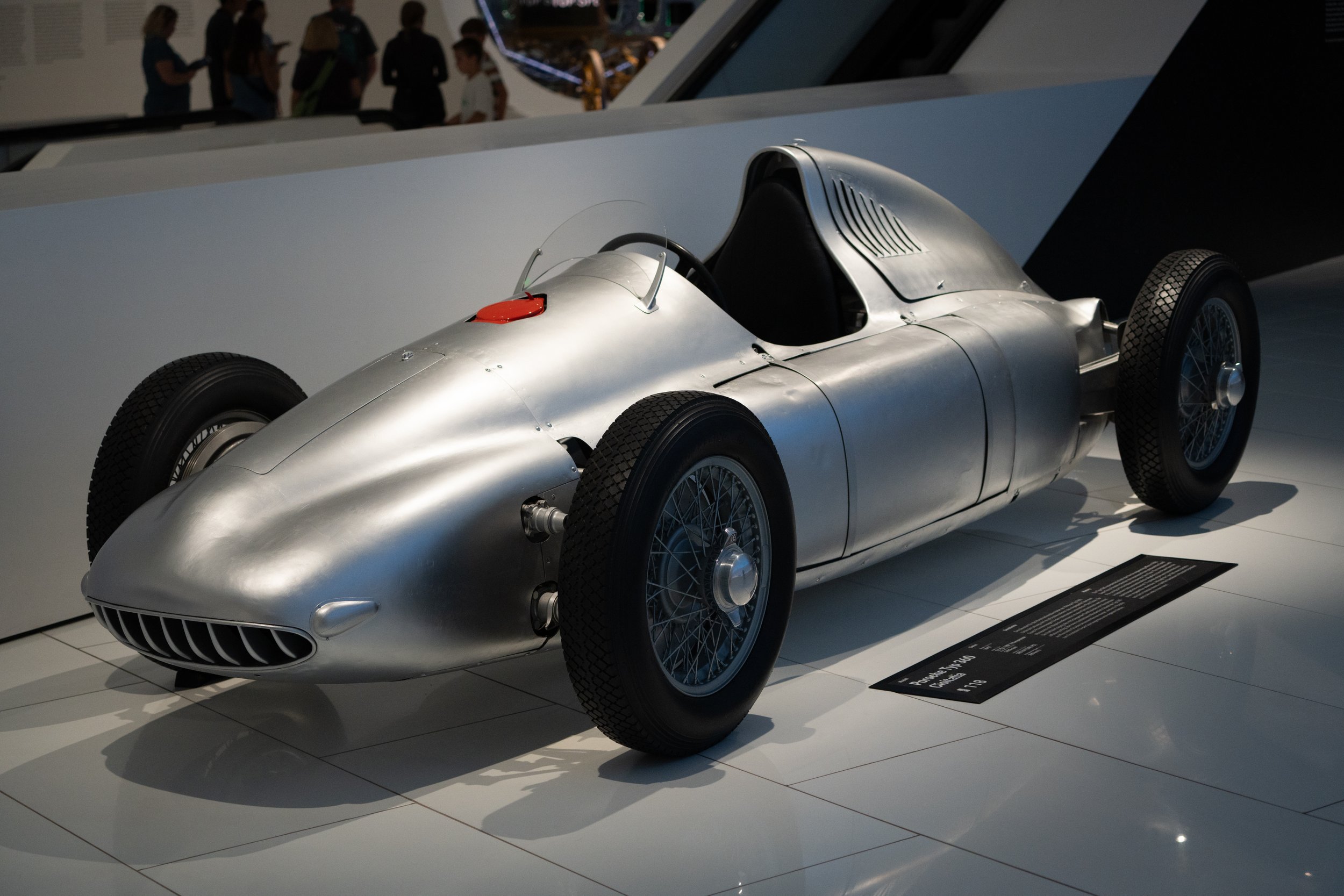 A vintage silver race car with an open cockpit and wire-spoke wheels on display in a museum, with a black informational plaque nearby.
