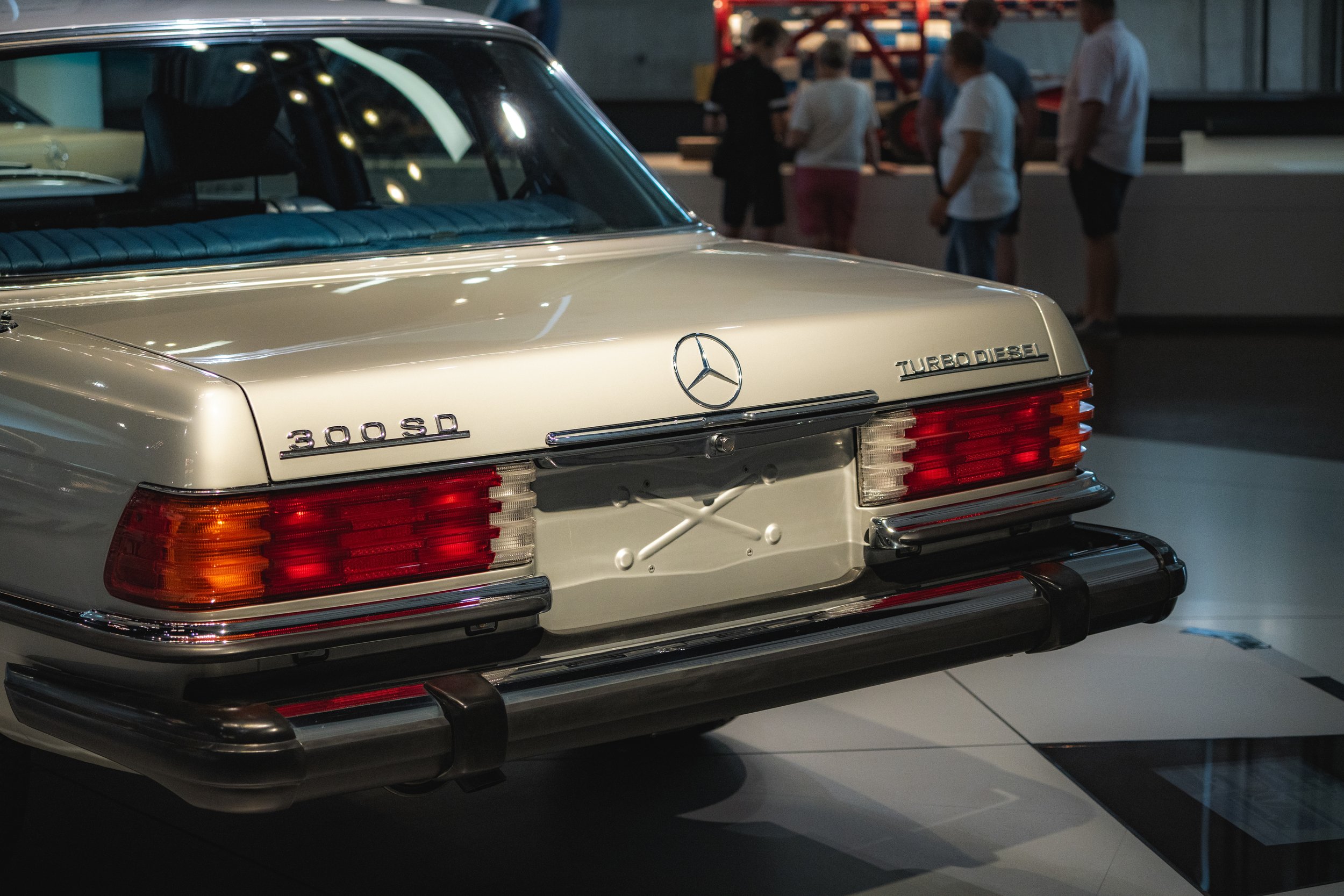 Rear view of a vintage Mercedes-Benz 300 Sd Turbo Diesel car displayed indoors, with some museum visitors in the background.