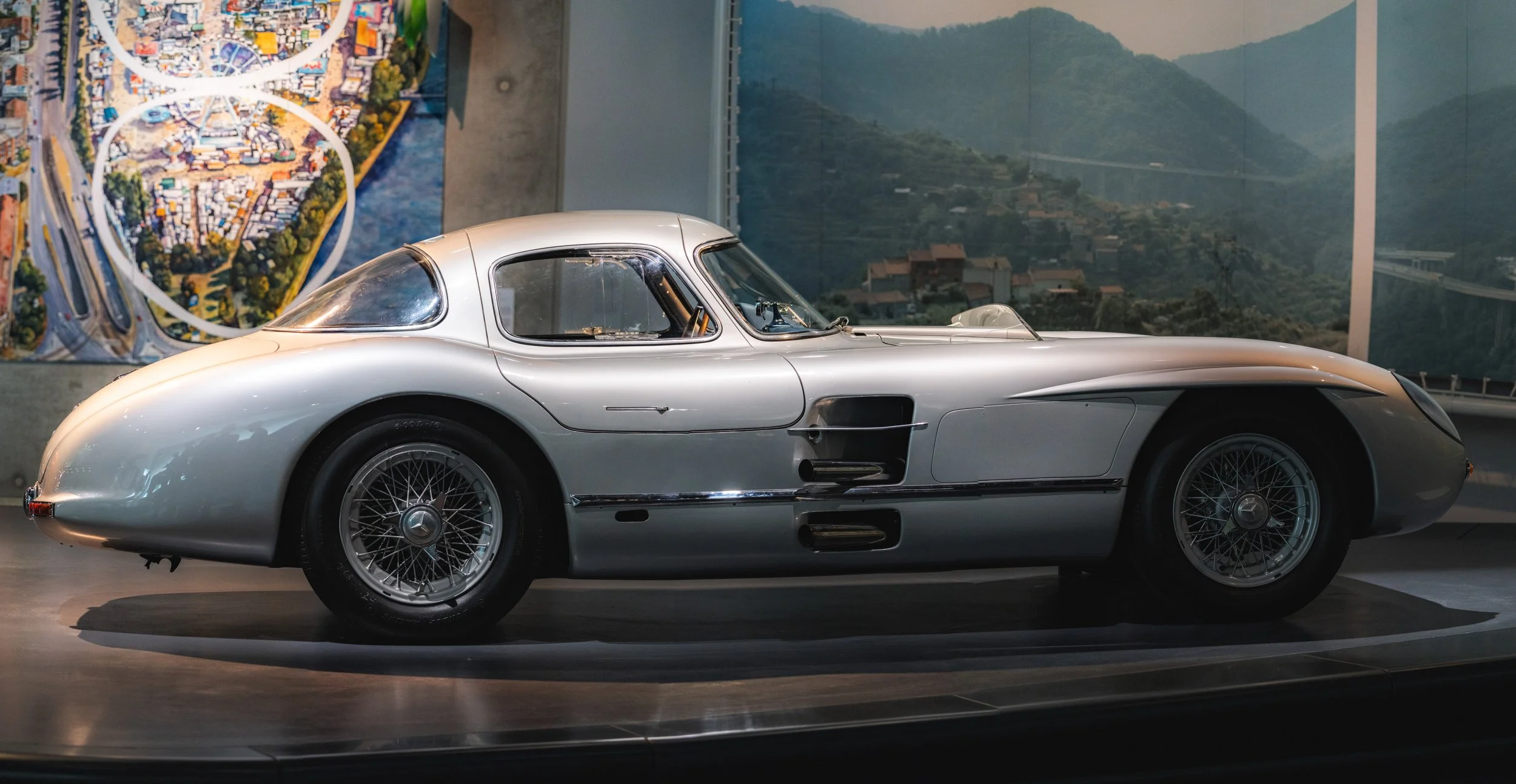 A silver vintage race car displayed in a museum with a mural and landscape photograph in the background.
