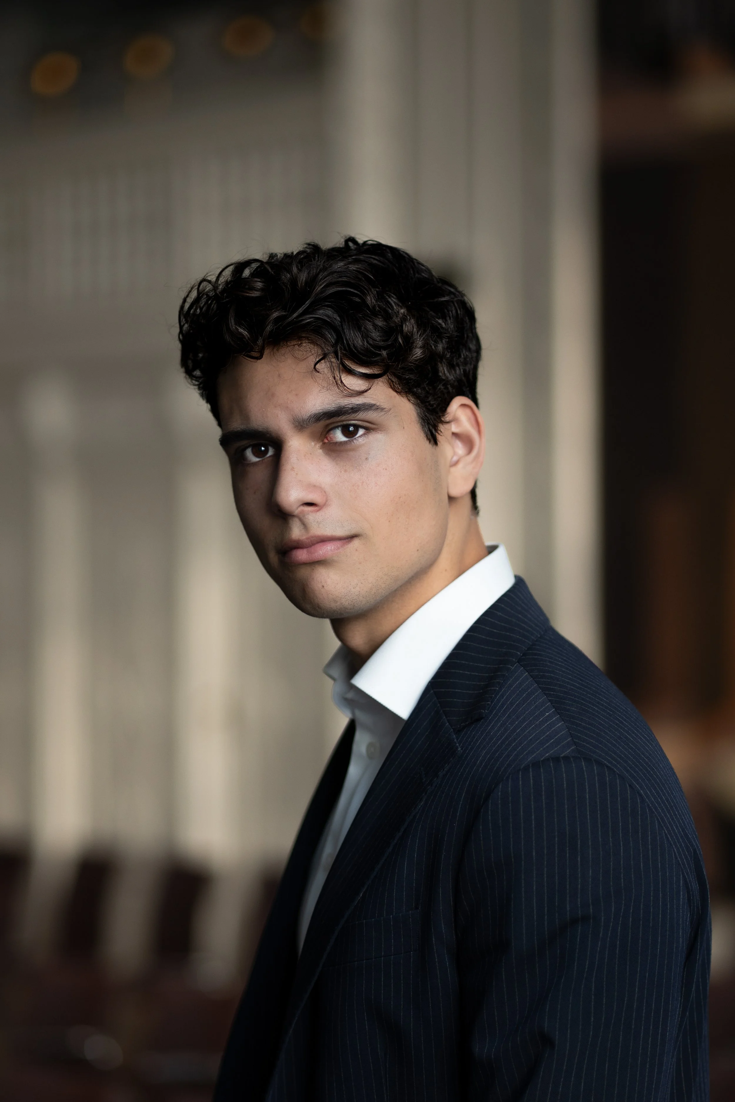 A young man with dark, curly hair and light skin, wearing a dark pinstripe suit and white shirt, looking at the camera with a neutral expression, standing indoors with blurred chairs and windows in the background.