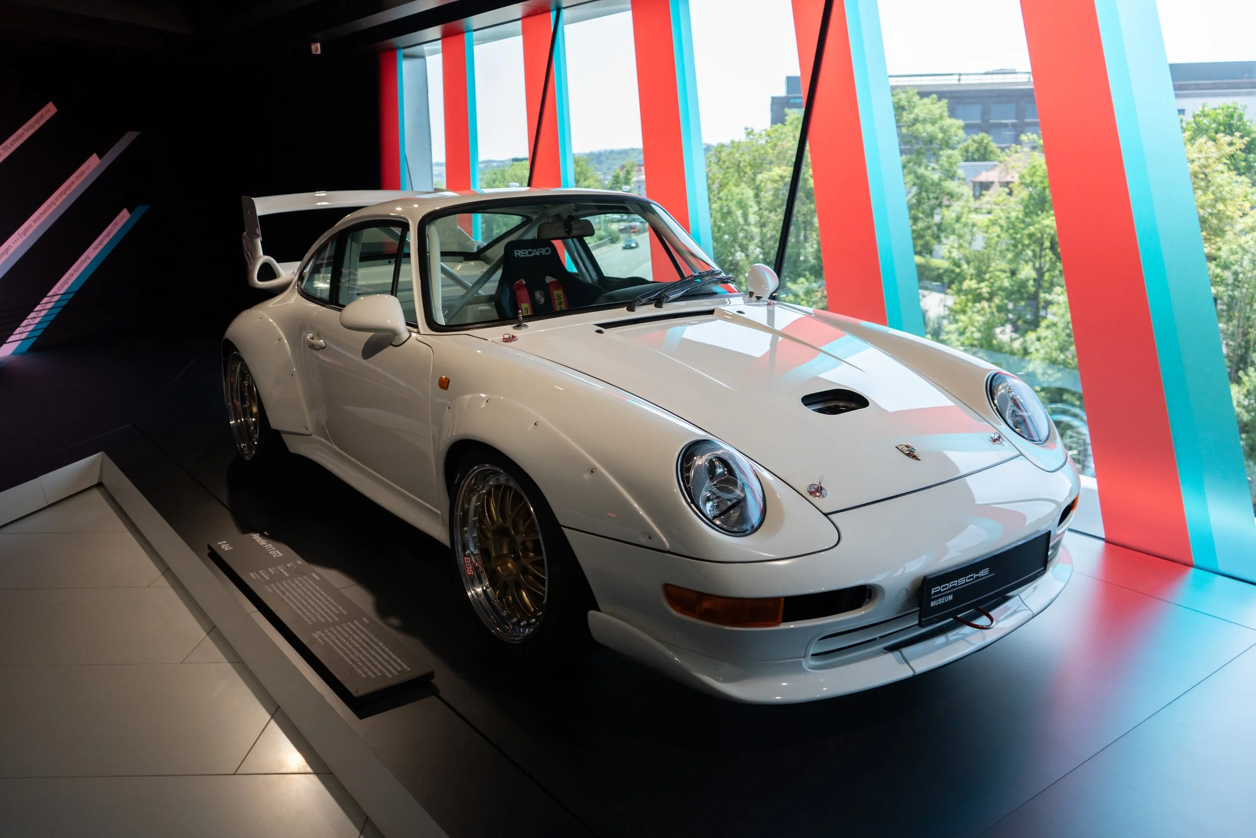 A white Porsche 911 racing car displayed in a museum with large windows and colorful vertical accents.