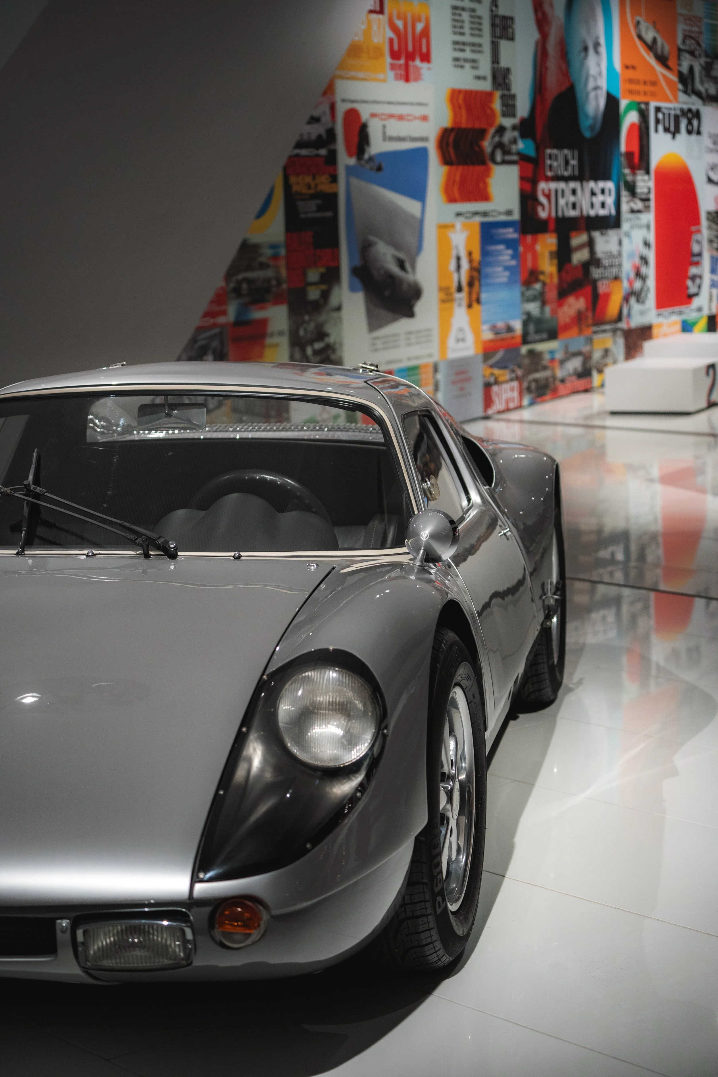 A silver vintage race car displayed in a museum, with colorful posters and magazine covers in the background.