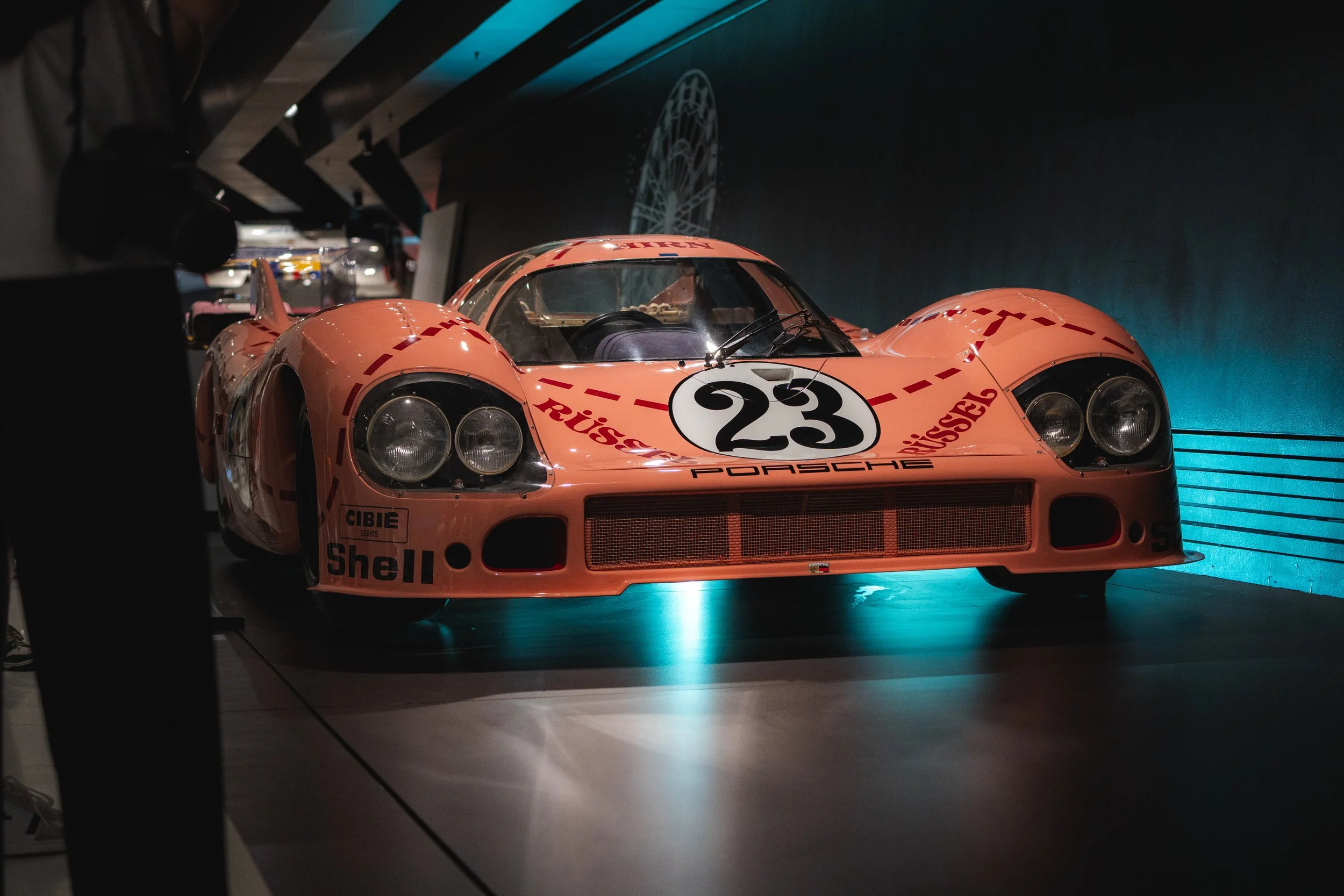A vintage Porsche race car, painted in pink with the number 23 on the hood, displayed in a dimly lit museum or exhibit, with branding from Shell and other sponsors.
