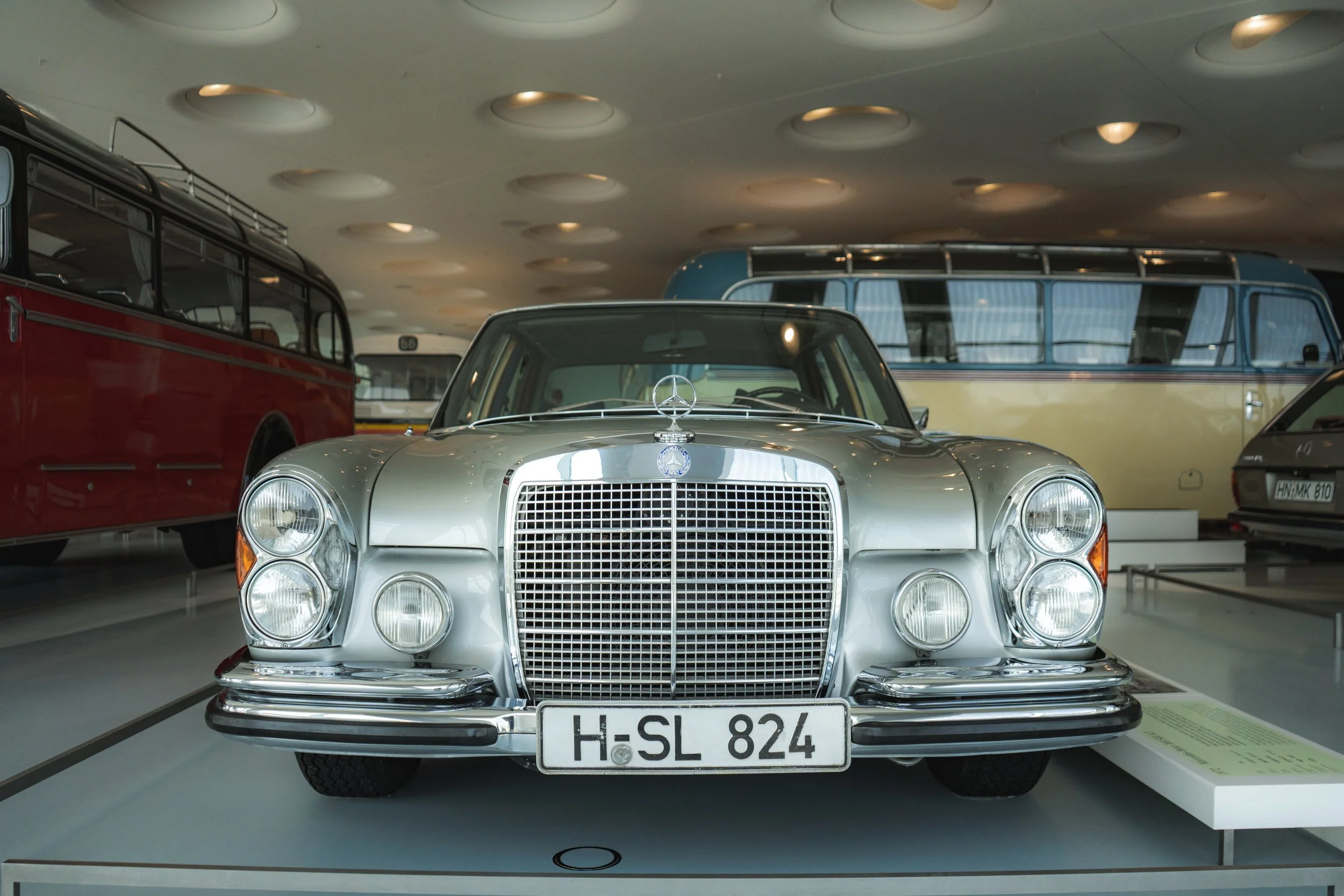 A silver vintage Mercedes-Benz car with a large front grille, four headlights, and the Mercedes logo on the hood, displayed in a museum with other classic vehicles in the background.