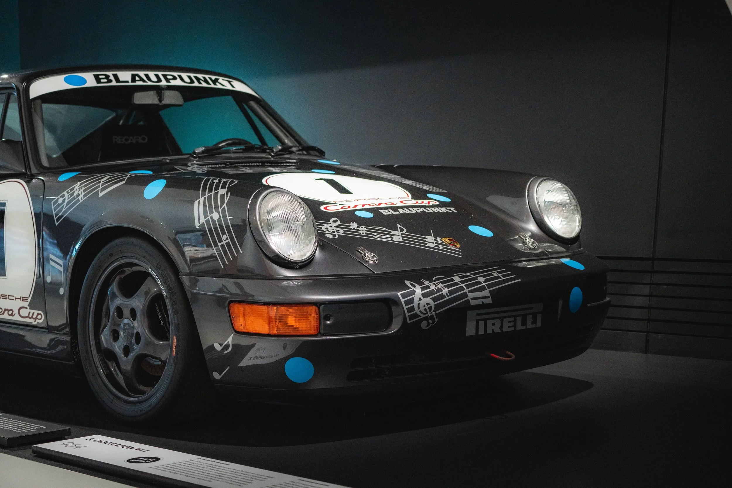A vintage Porsche race car with a dark gray body decorated with musical notes and blue dots, displayed in a museum.