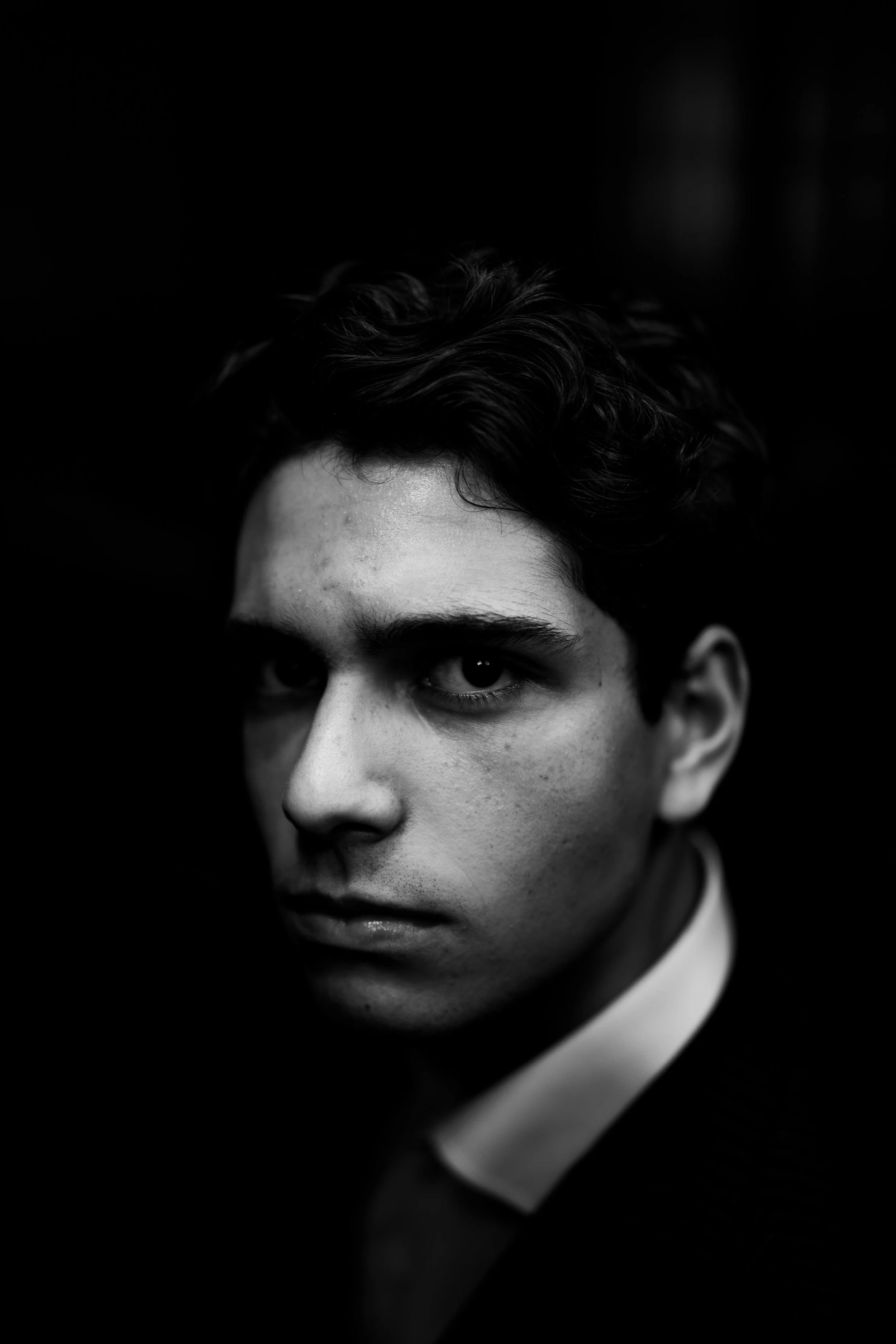 Black and white portrait of a young man with dark hair, looking intensely at the camera, with shadows covering part of his face.