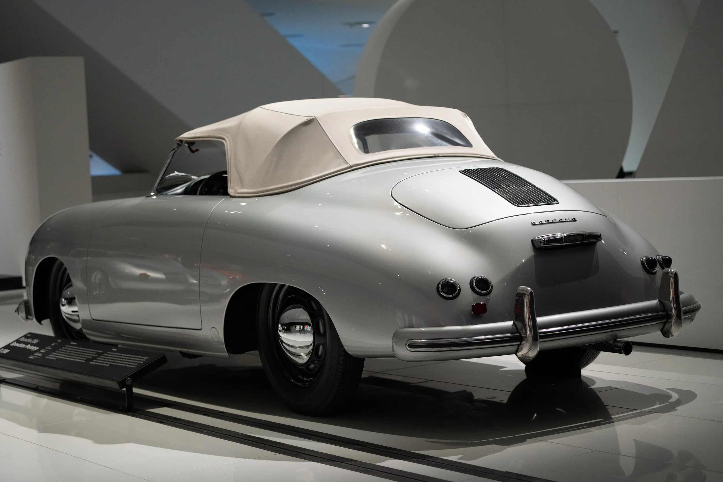 A vintage silver Porsche 356 convertible with a beige soft top on display in a museum.