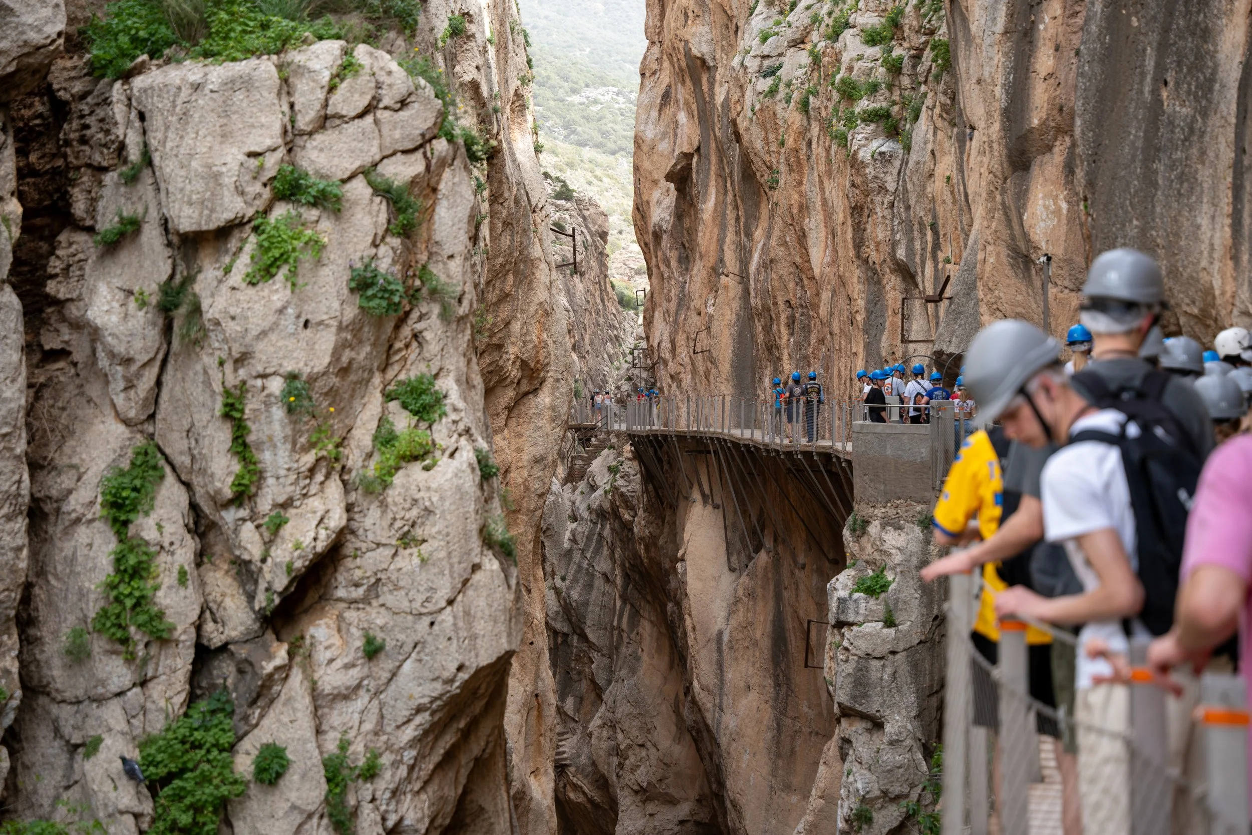 Tourists wearing helmets and backpacks walk along a narrow mountain trail with a metal railing, passing through a deep rocky canyon with steep cliffs on either side and some green vegetation.
