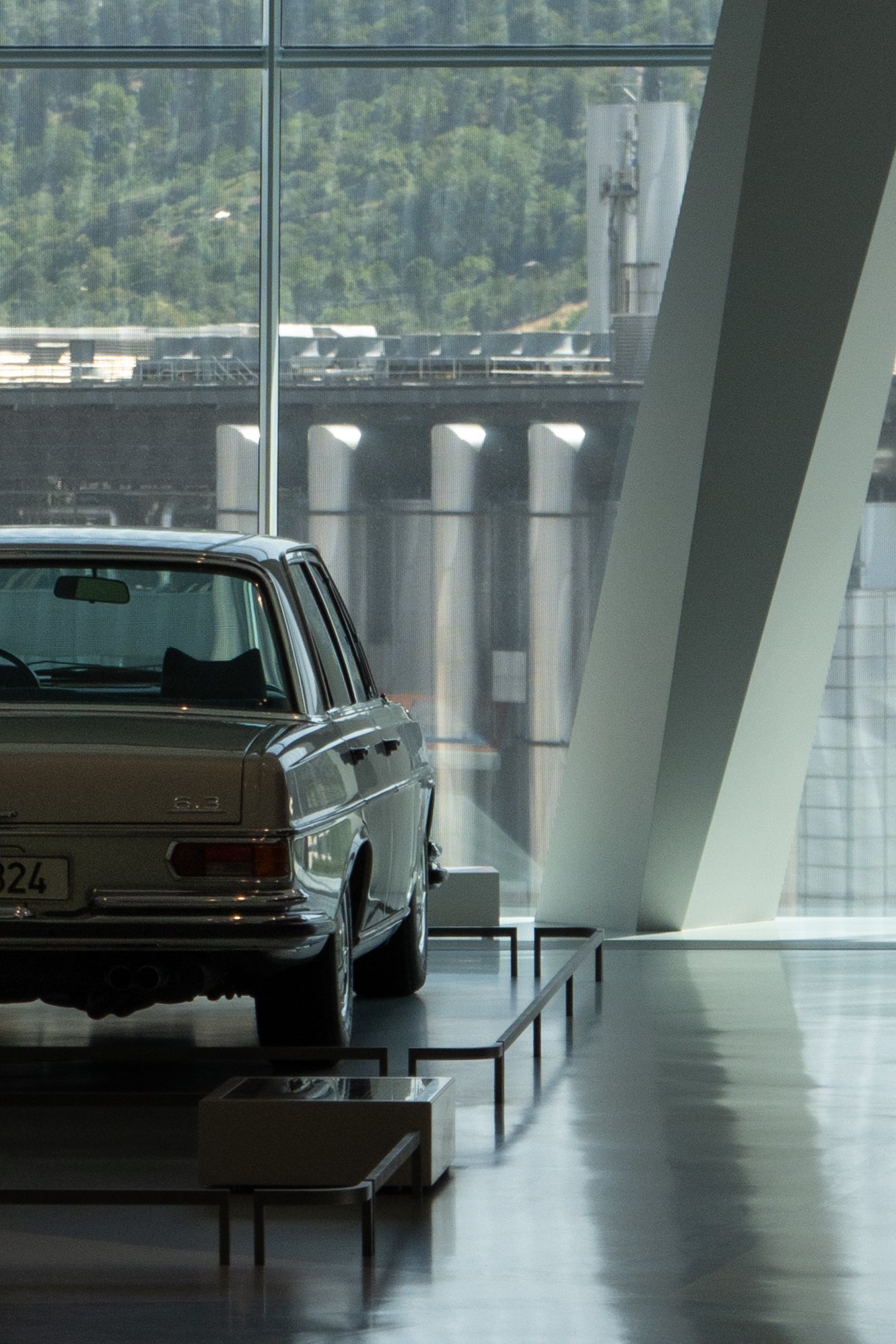 A vintage silver car displayed inside a modern building with large windows and a view of an industrial bridge and green hills outside.