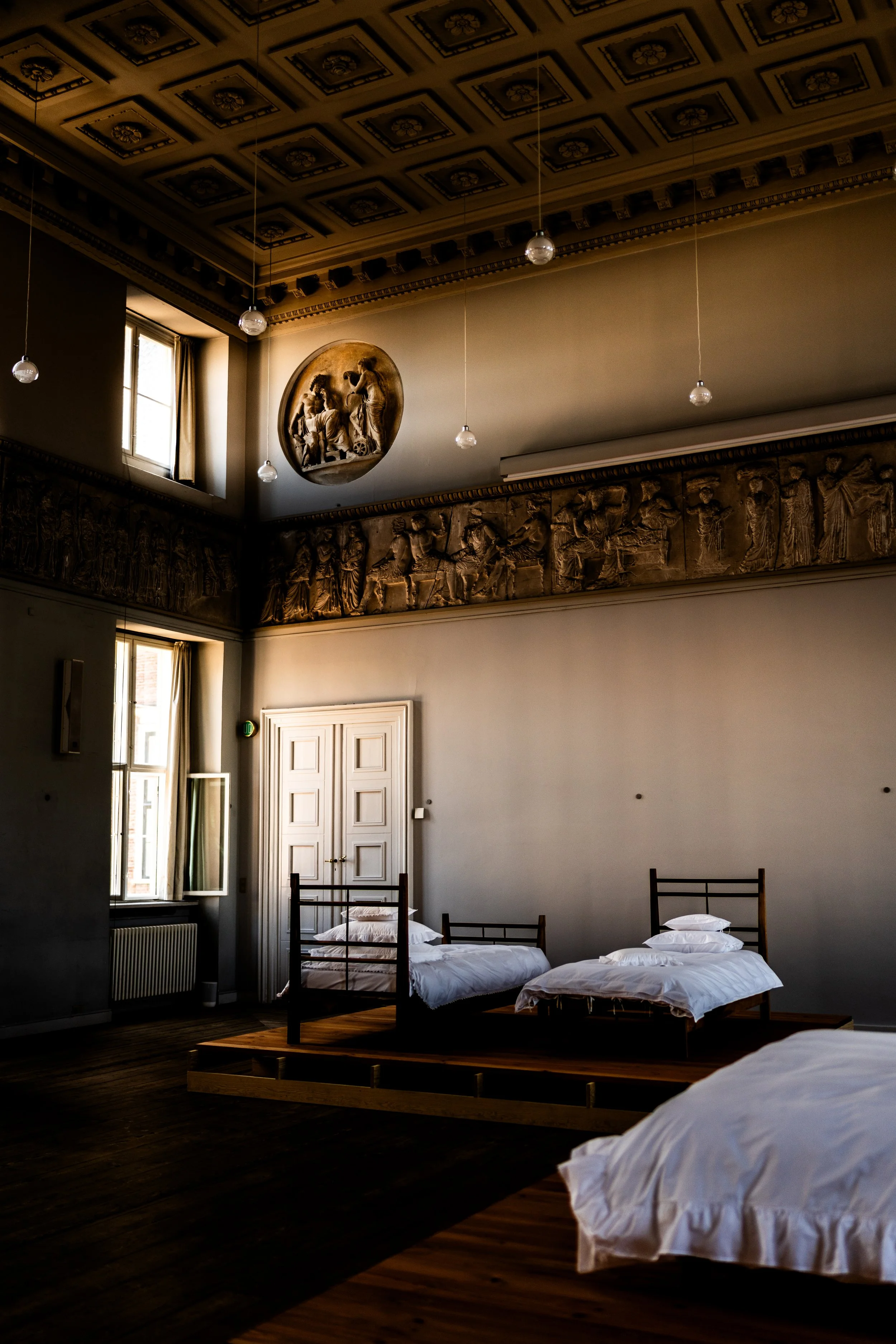 A room with three beds with white bedding, wooden bed frames, and a high ceiling with decorative molding and an ornate frieze. There are tall windows letting in natural light, and historical relief sculptures on the upper walls.